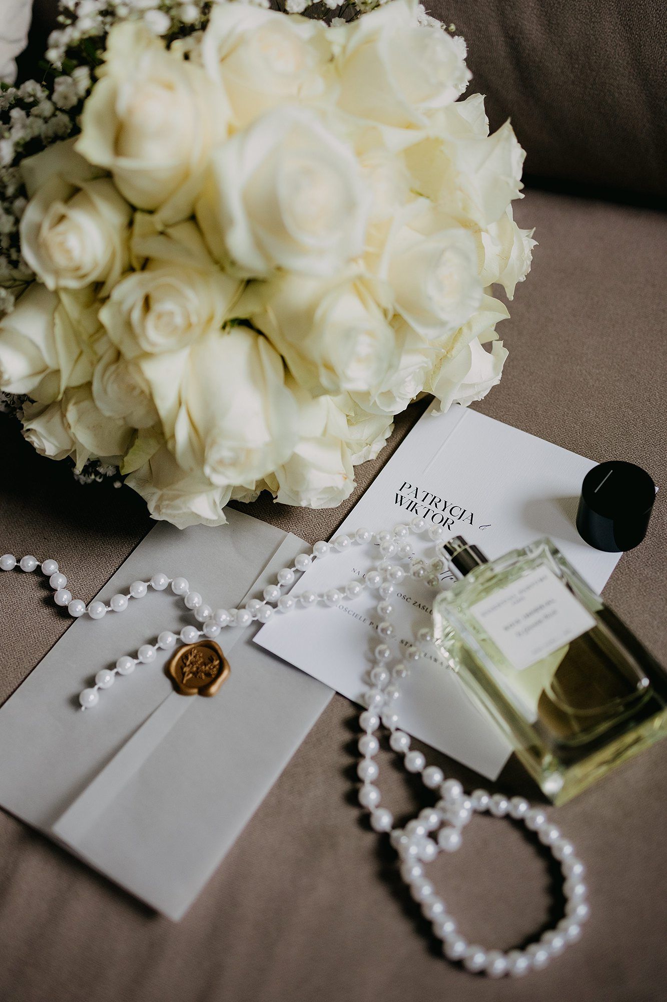Elegant bridal flat lay featuring white roses bouquet, pearl necklace, perfume bottle, and wedding invitation.