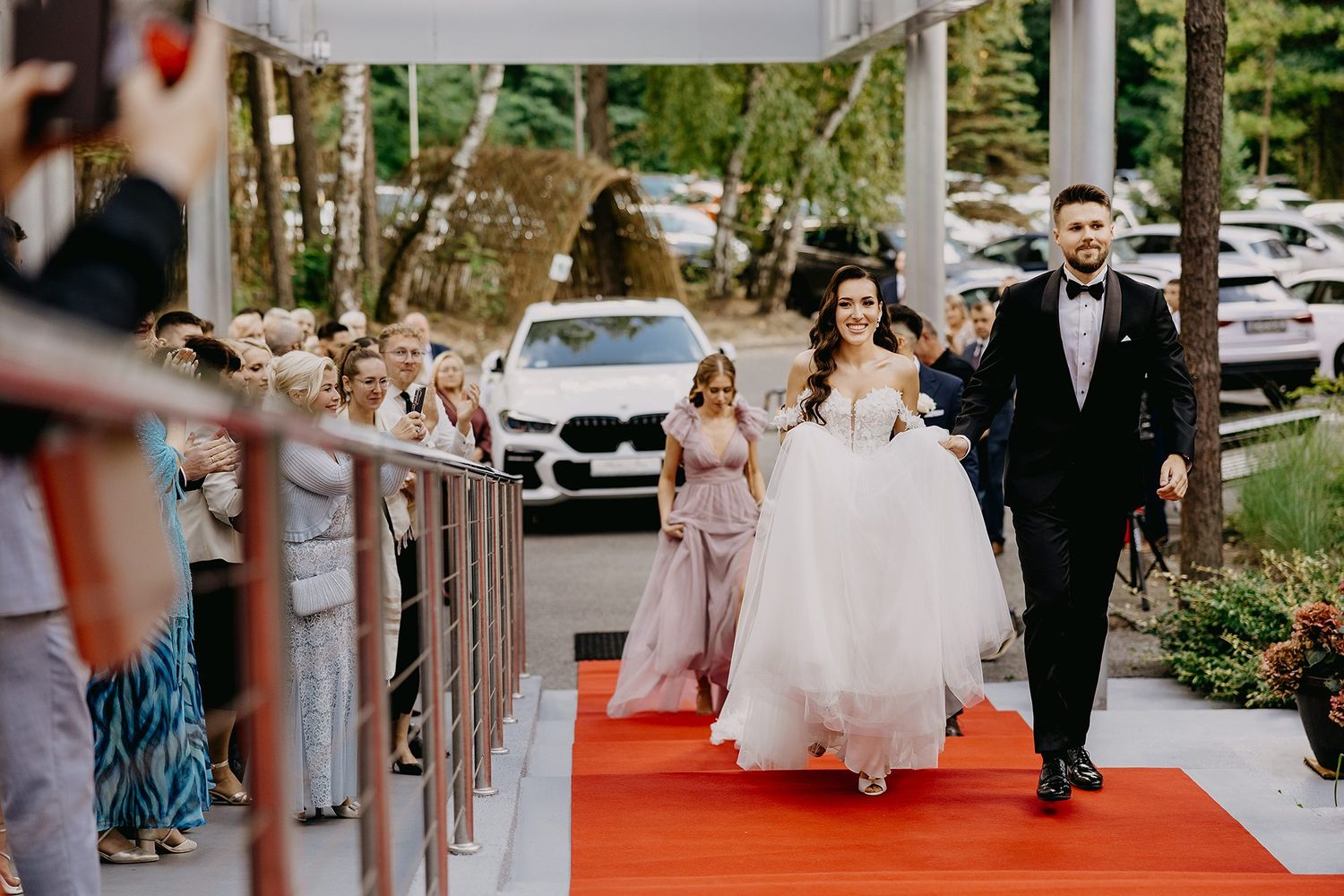 Bride and groom walking down red carpet aisle surrounded by wedding guests at outdoor ceremony.