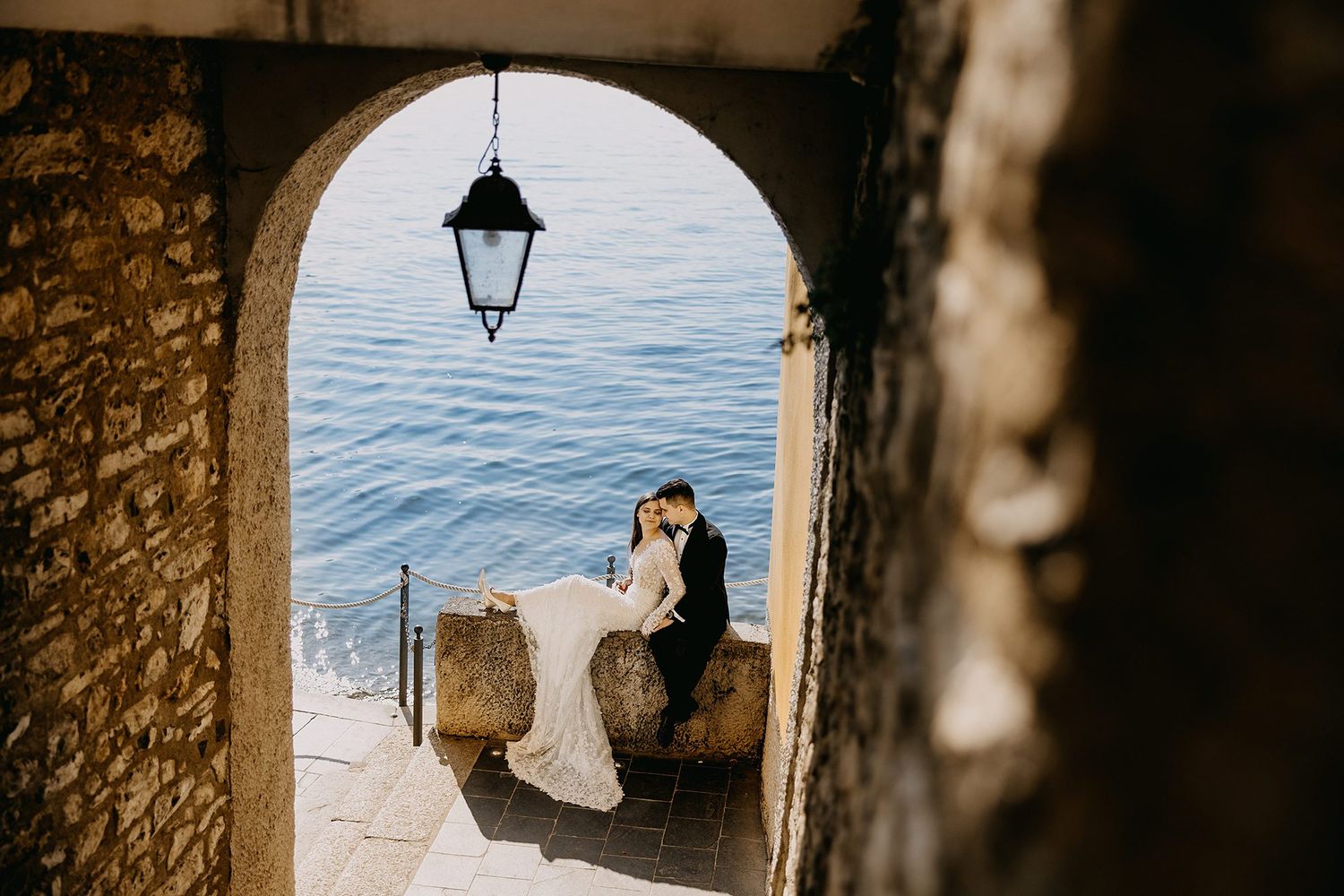 Newlyweds share intimate moment seated by the water framed by a rustic stone archway with hanging lantern.