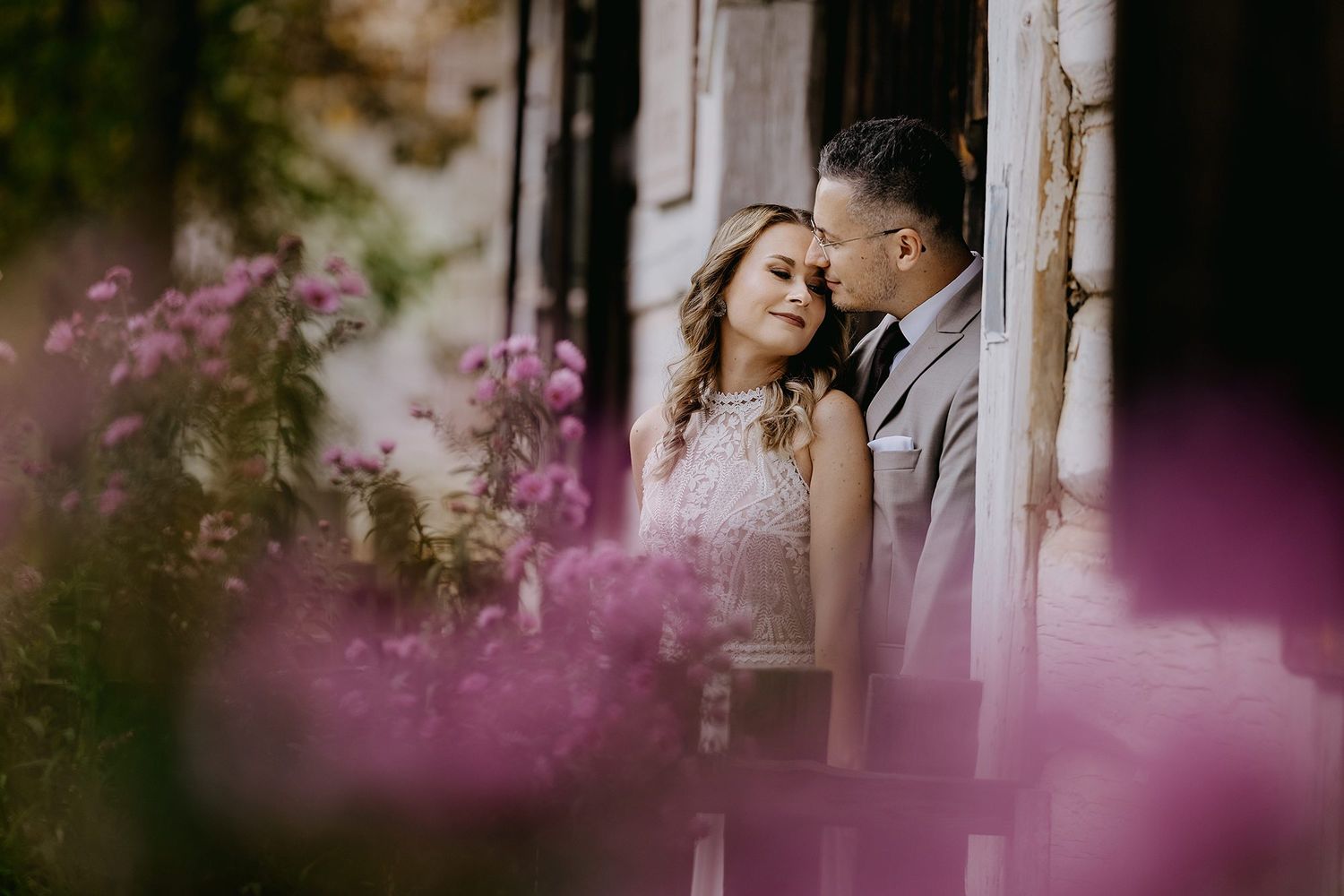 Couple sharing an intimate moment framed by vibrant pink flowers near a rustic doorway.
