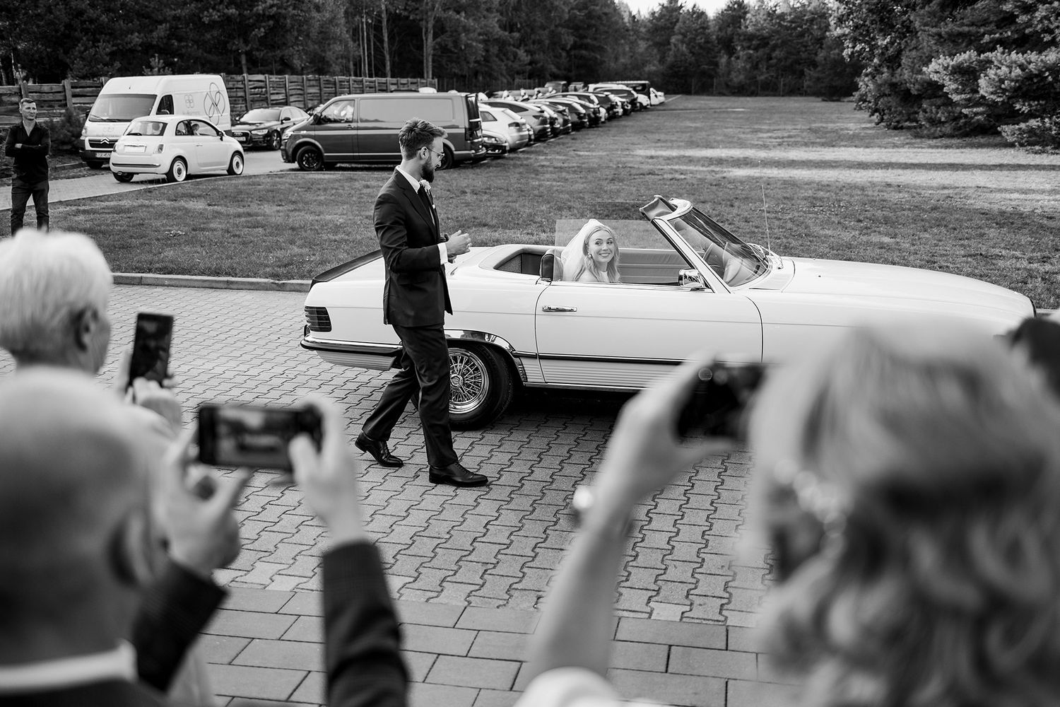Black and white photo of bride in white convertible classic car with wedding guests taking photos.