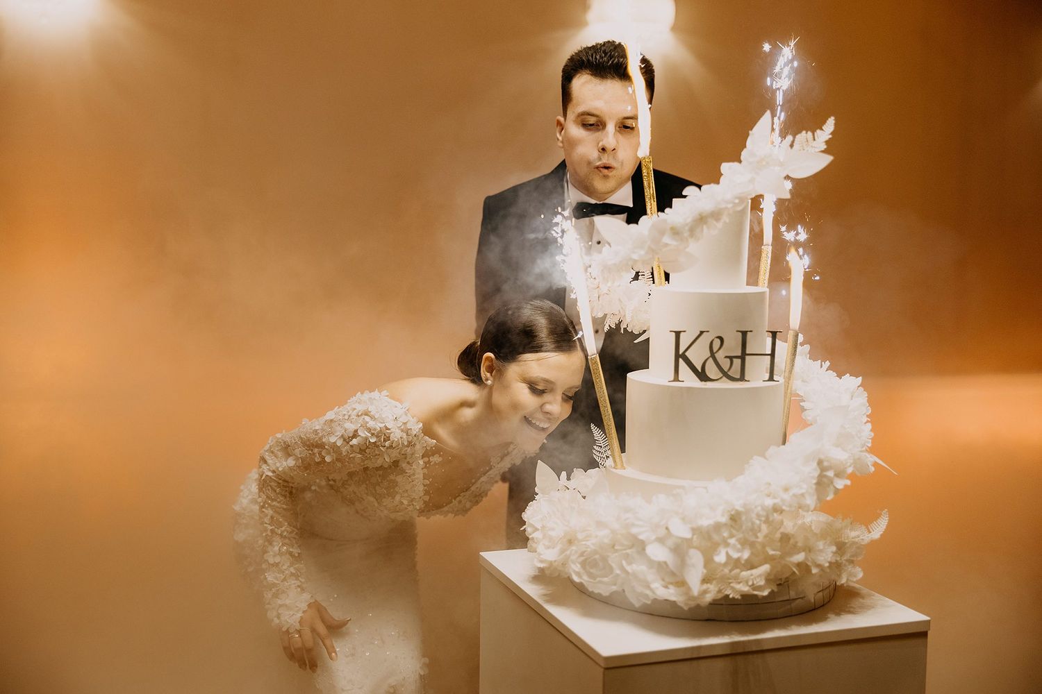 A bride and groom cut their elegant white wedding cake monogrammed with K&H amid golden smoke and sparklers.
