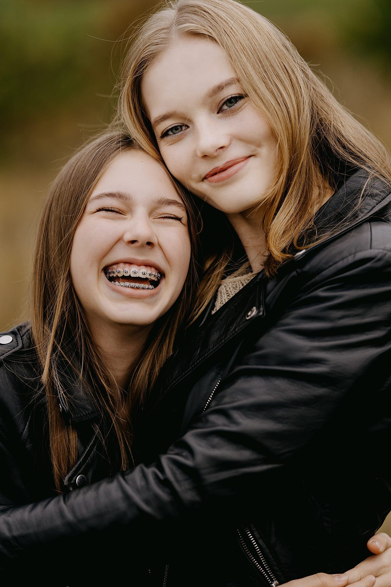 Two teenage girls laughing and hugging wearing black leather jackets outdoors.