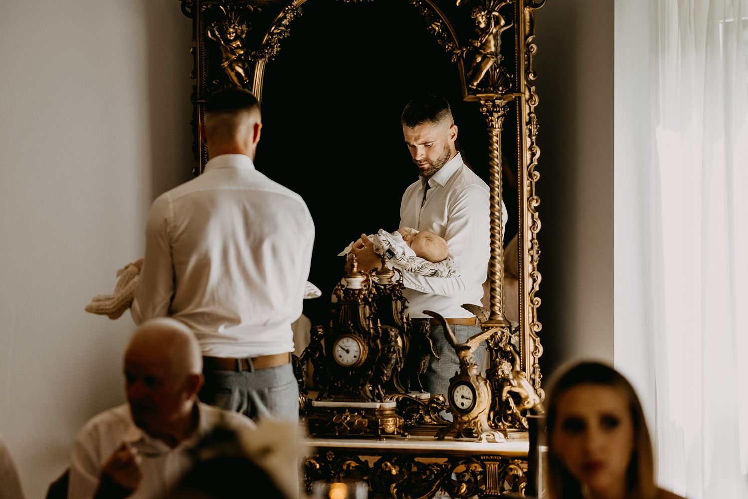 Man in white shirt adjusts clothing while reflected in an ornate gold-framed mirror during a formal event.