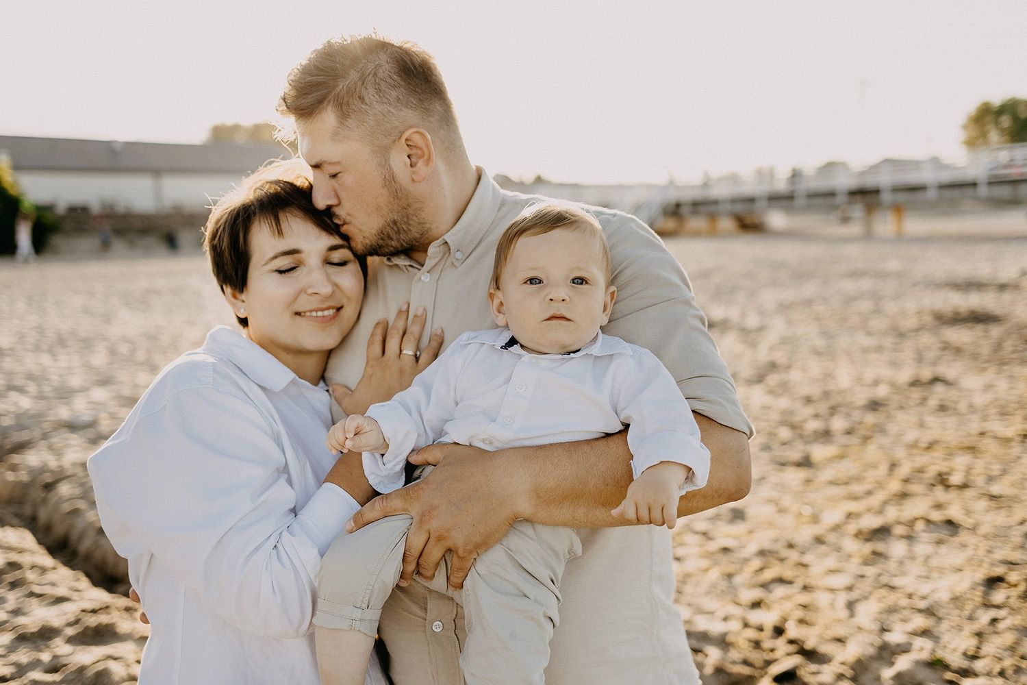 Young couple with baby boy shares a tender kiss on a sandy beach.