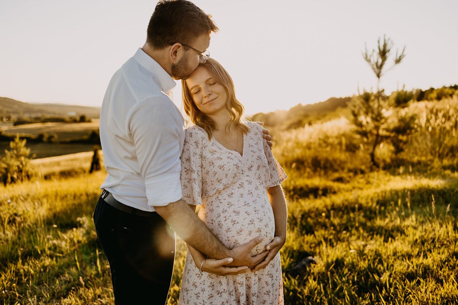 Couple embracing in golden sunset meadow, pregnant woman in floral dress cradling her baby bump.