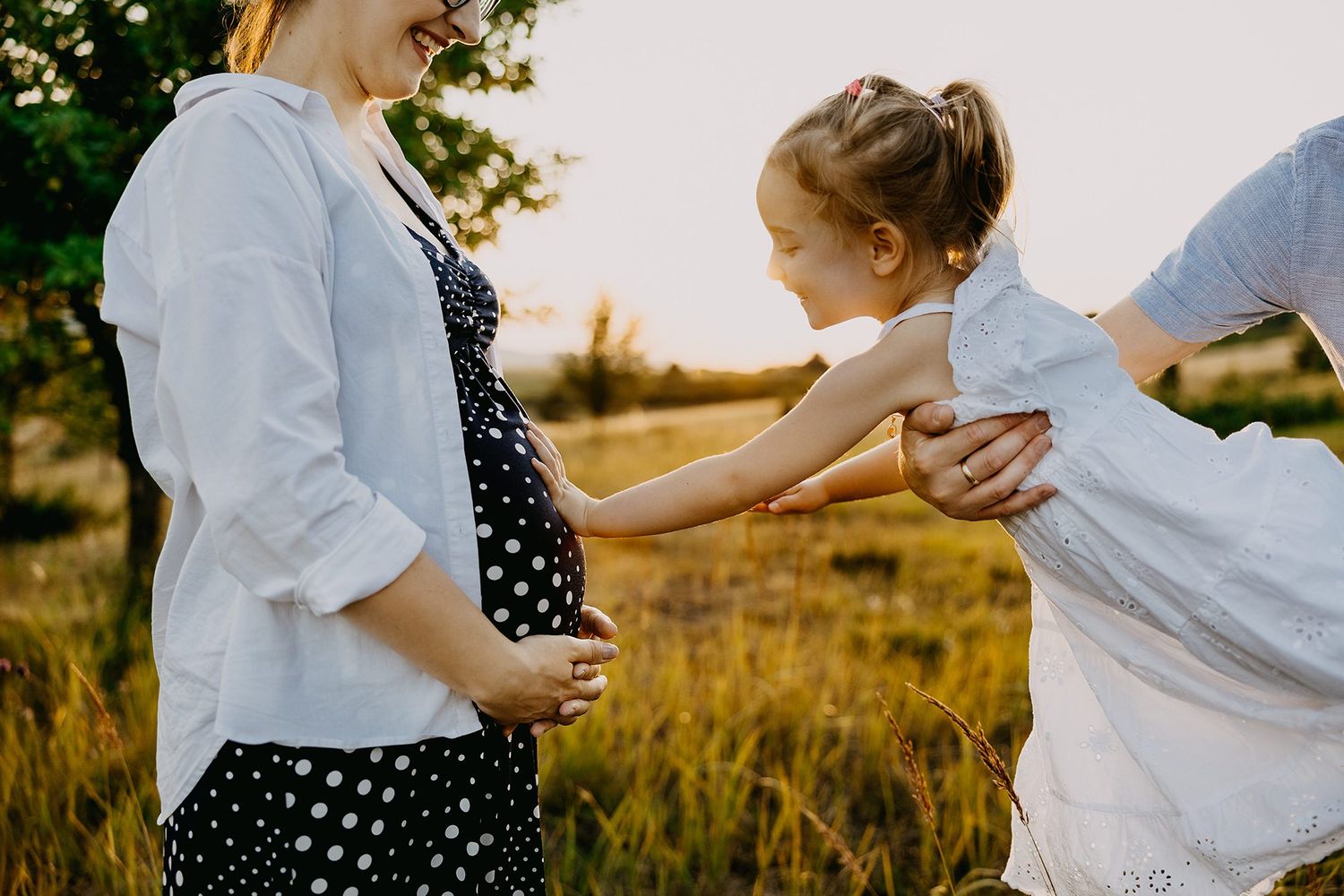 A pregnant woman and child hold hands in a sunlit meadow during a family photo session.
