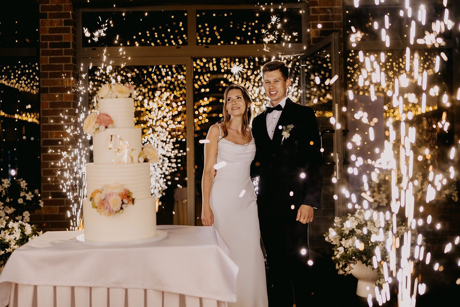 Bride and groom cutting wedding cake surrounded by sparkling fireworks at elegant indoor wedding reception.