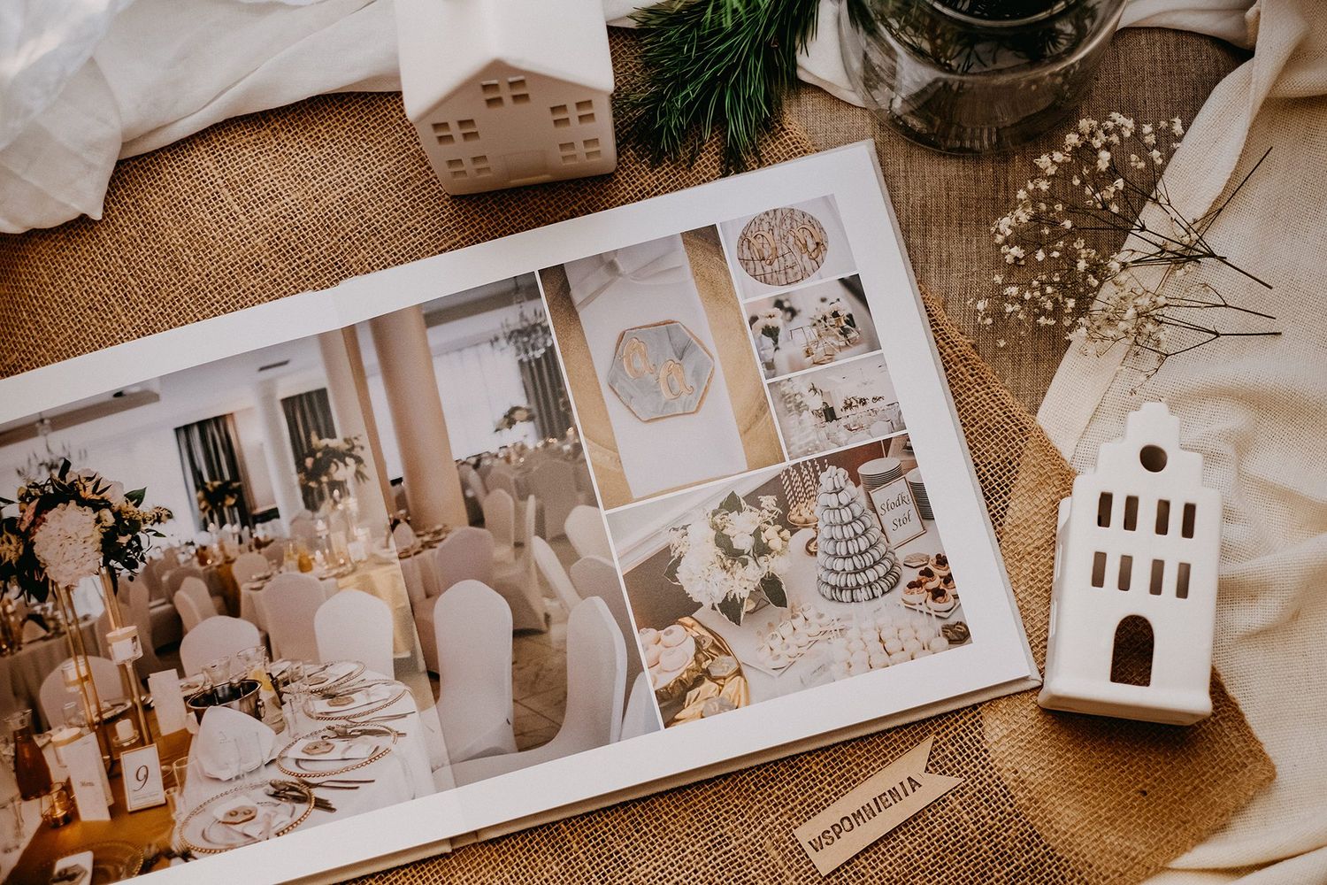 Open wedding photo album displaying elegant reception decor on burlap with decorative house figurines and dried flowers.