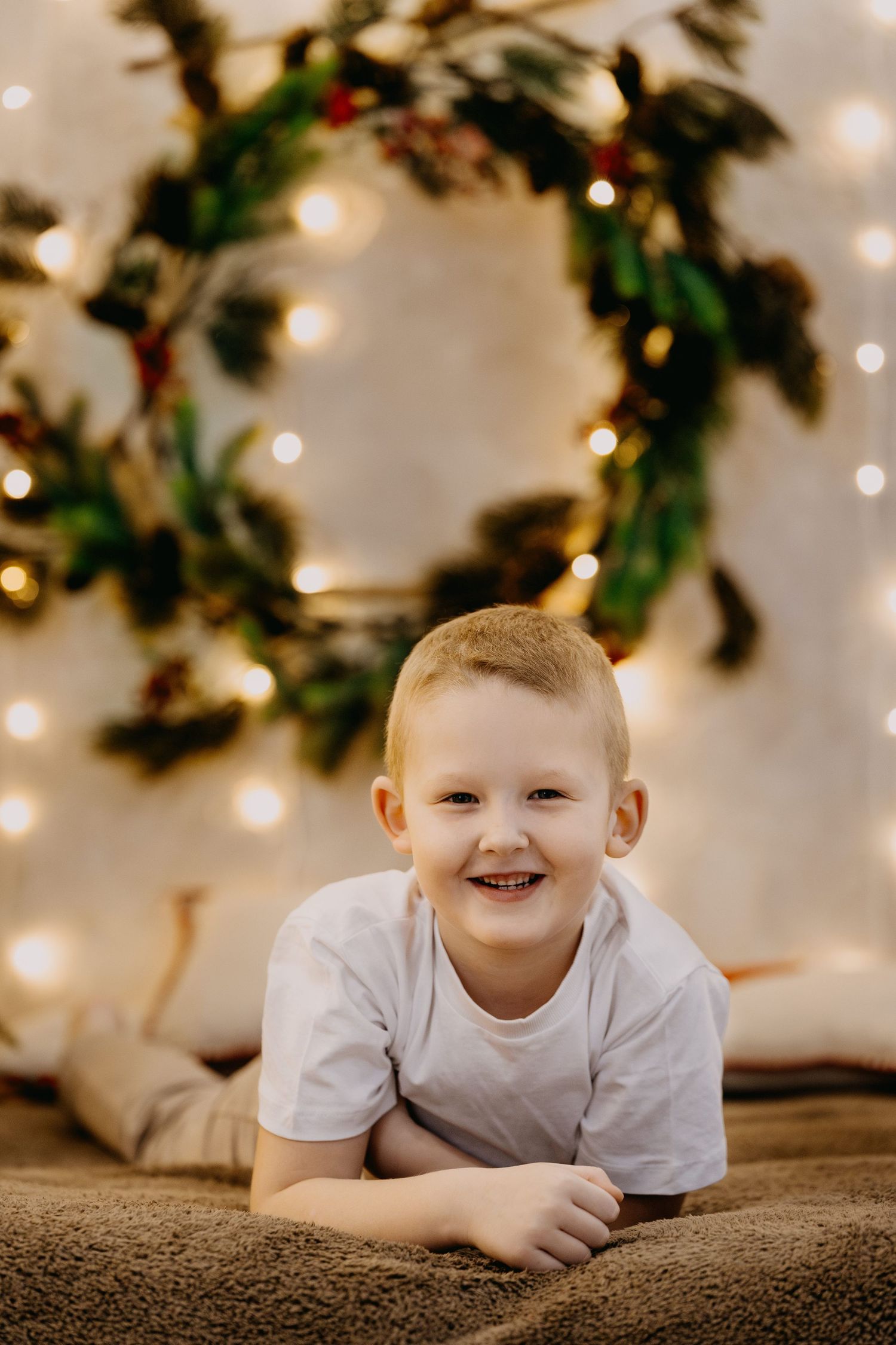Smiling blond boy in a white t-shirt lying on a soft brown blanket in front of a festive greenery wreath and warm twinkling lights.