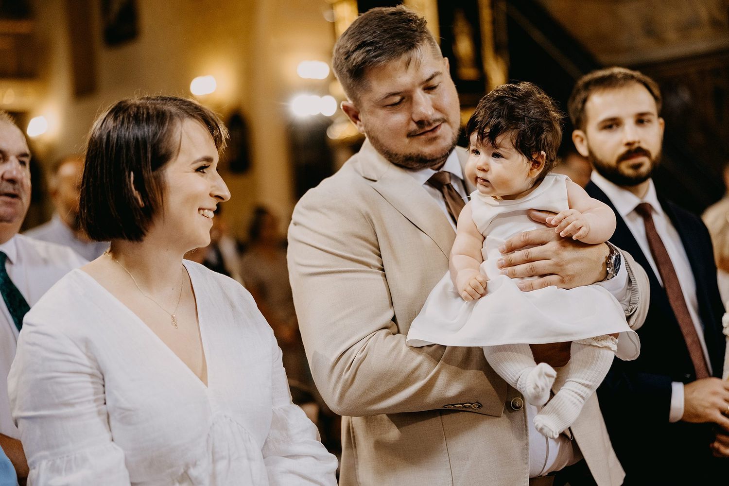 Smiling couple holding baby in white outfit during christening celebration in church.