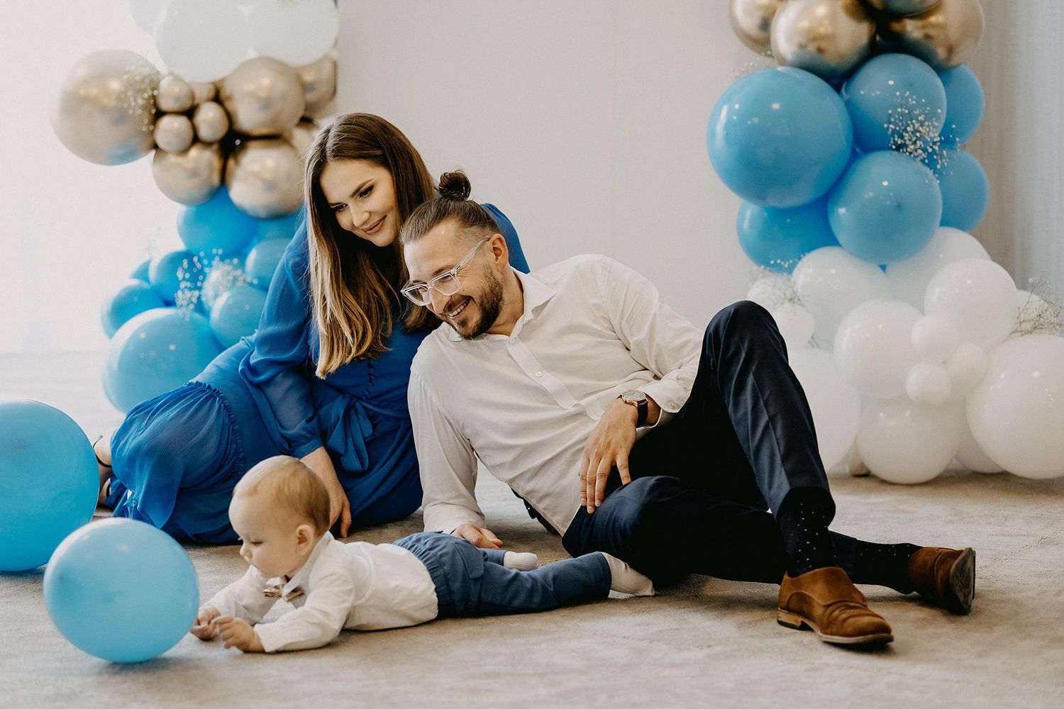 Happy parents sit on floor with baby surrounded by blue and white balloon garland decoration.