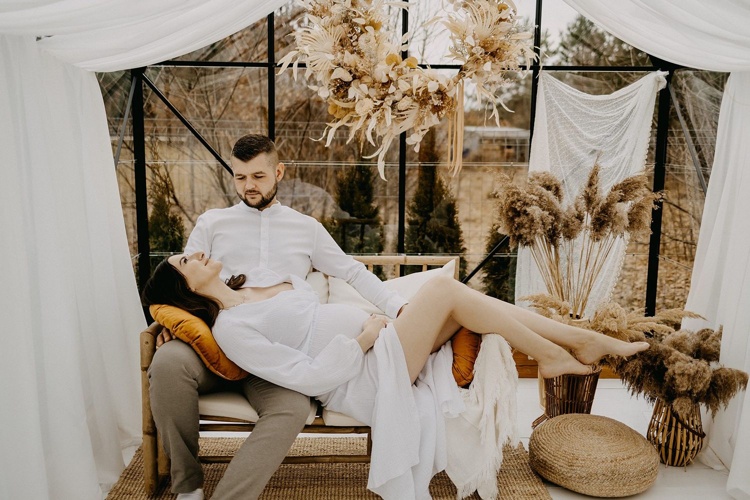 Couple relaxing together on bench in boho-decorated studio with dried flowers and white drapes.