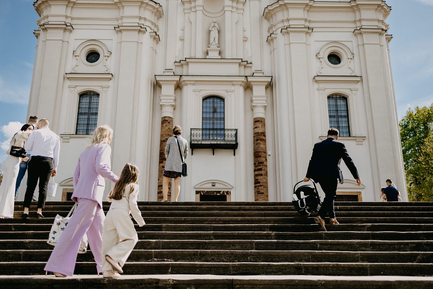 Family and guests climbing steps of grand white baroque church for baby baptism ceremony.