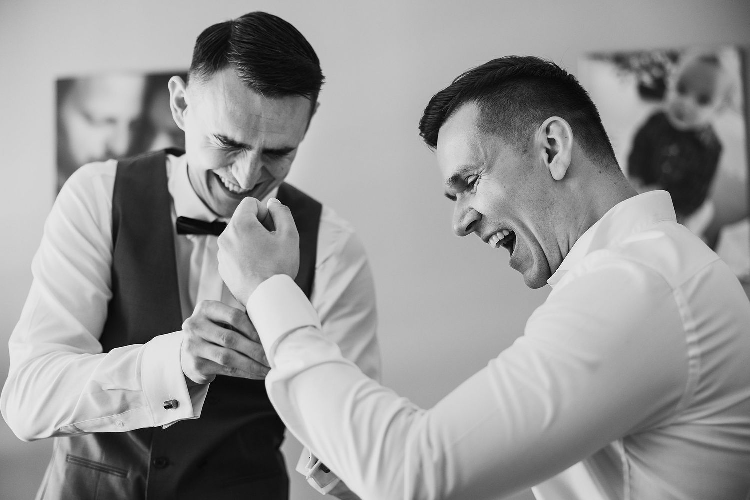 Black and white photo of groom laughing with groomsman while adjusting cufflinks before wedding ceremony.