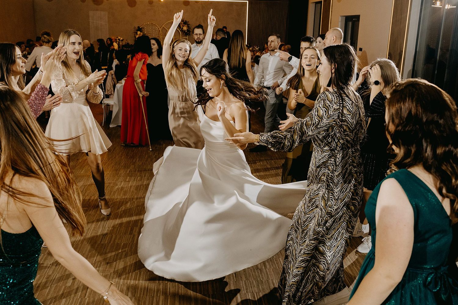 Bride twirls in white gown surrounded by joyful guests dancing at lively wedding reception party.
