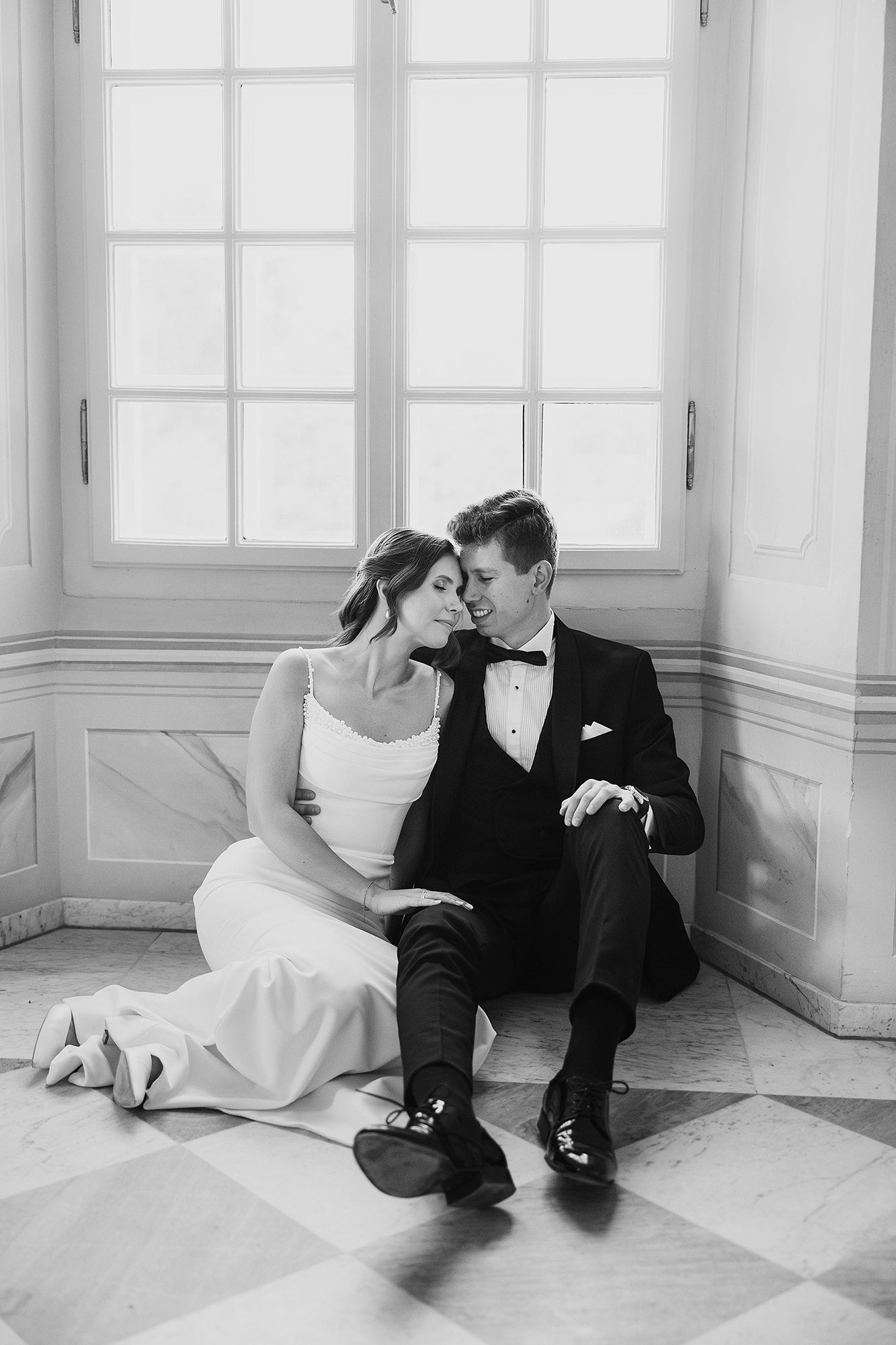 Black and white photo of a bride and groom sitting together on the floor by a large window.