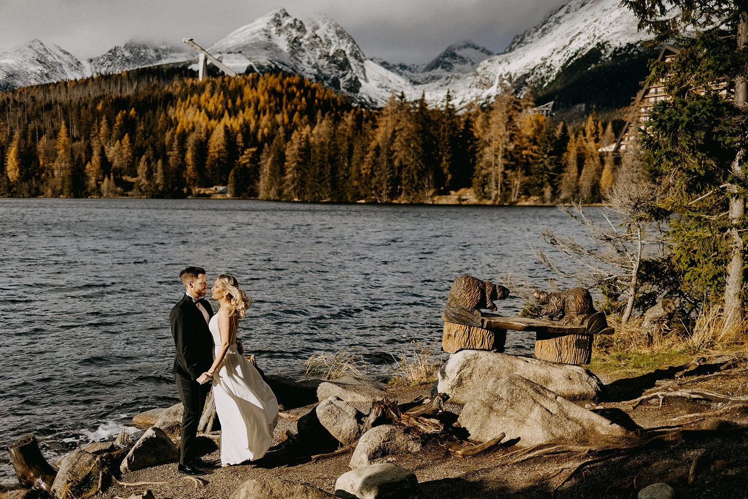 Wedding couple embraces on rocky lakeshore with snow-capped mountains and golden autumn trees.
