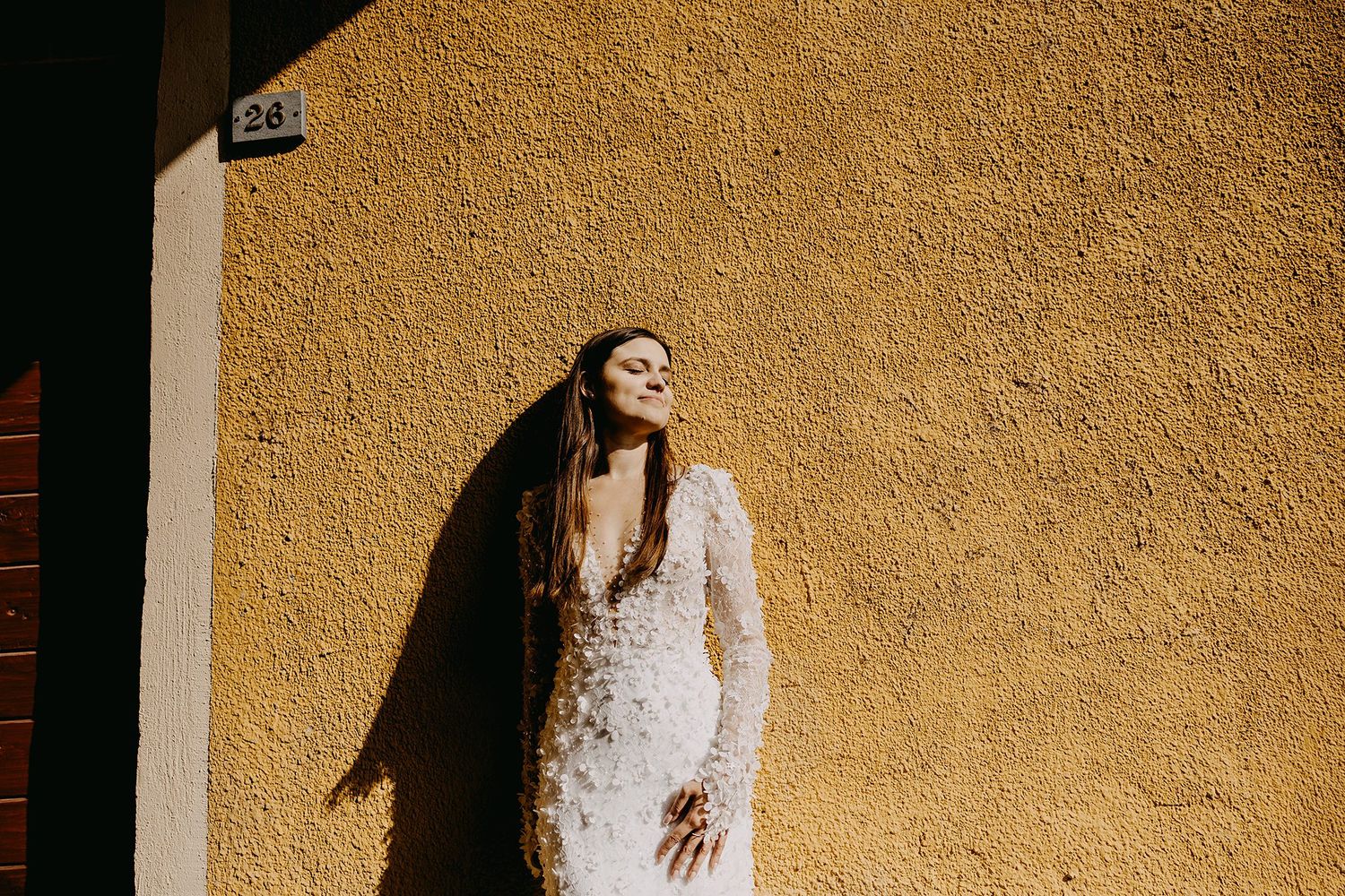 Radiant bride in long-sleeve lace wedding gown poses against textured yellow sunlit wall.
