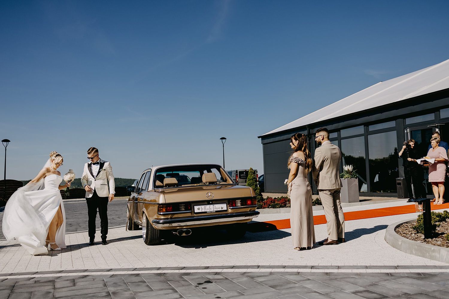 Wedding couple arriving in a vintage gold car at a modern venue under a clear blue sky.