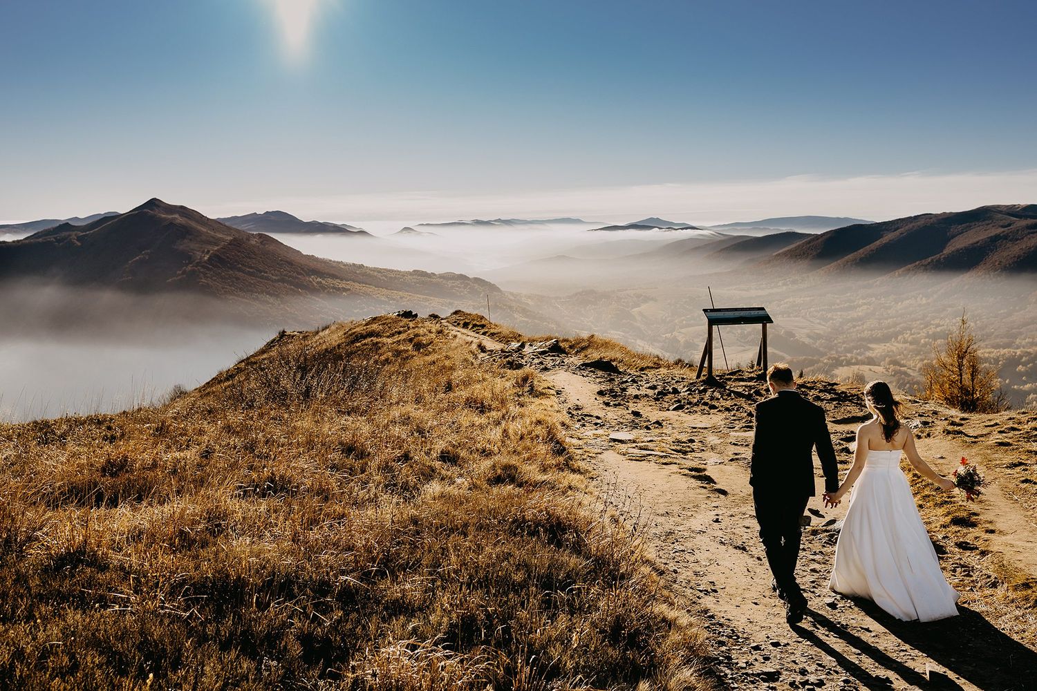Wedding couple holding hands on a scenic mountain path with clouds and rolling hills in the background.