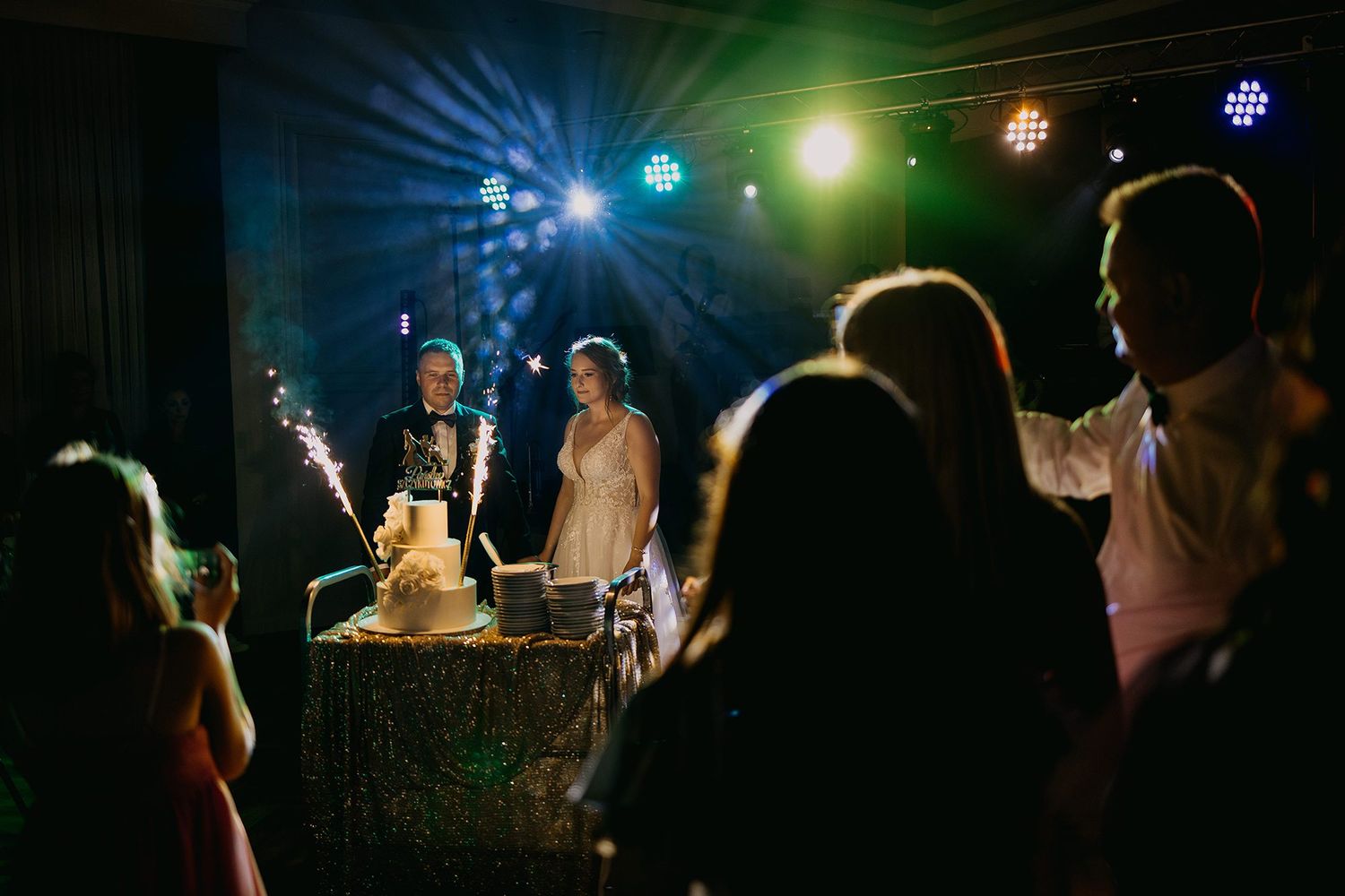 Bride and groom cutting wedding cake with sparklers under colorful disco lights on the dance floor.