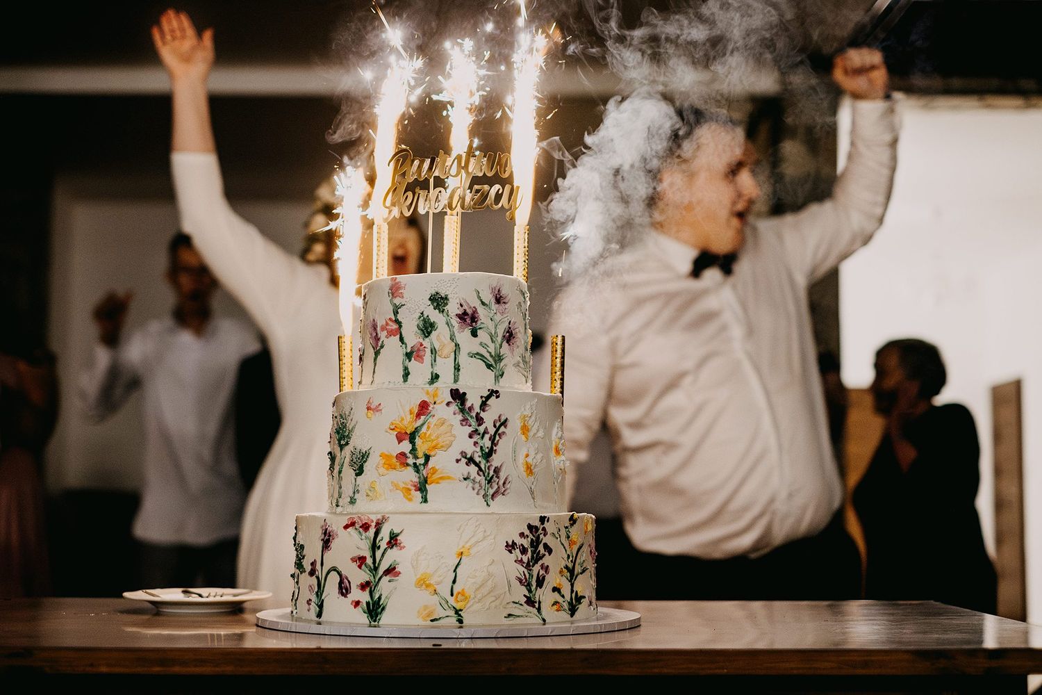 Three-tier floral wedding cake with gold candles and sparklers as guests celebrate joyfully in the background.