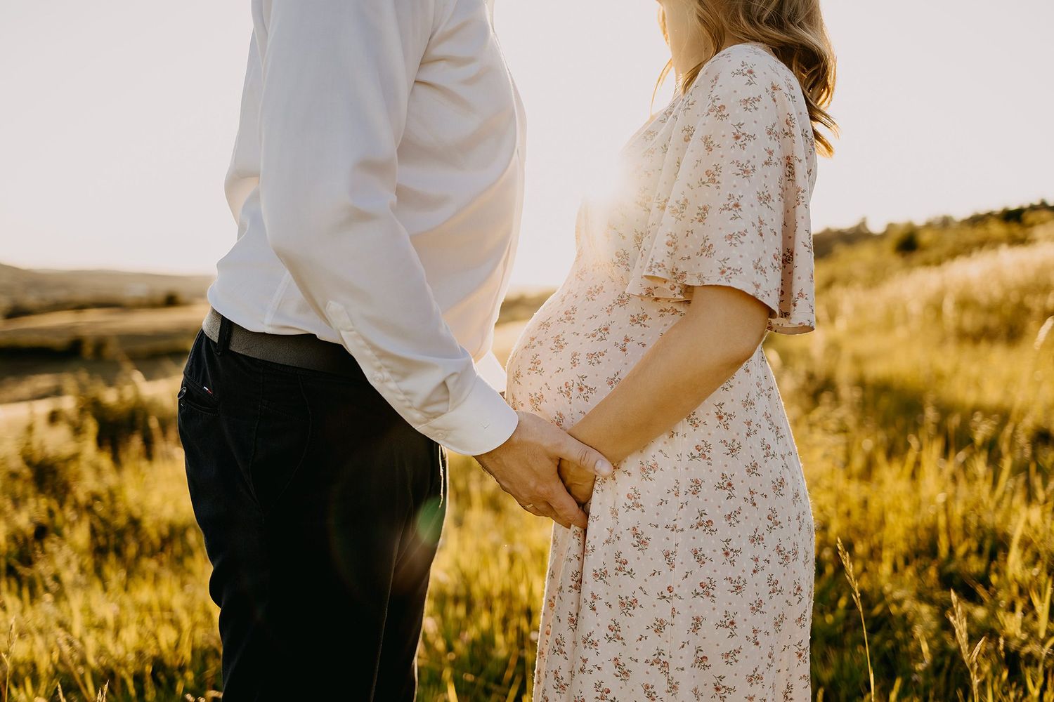 Pregnant couple holding hands in a sun-drenched meadow, woman in floral dress cradling baby bump.