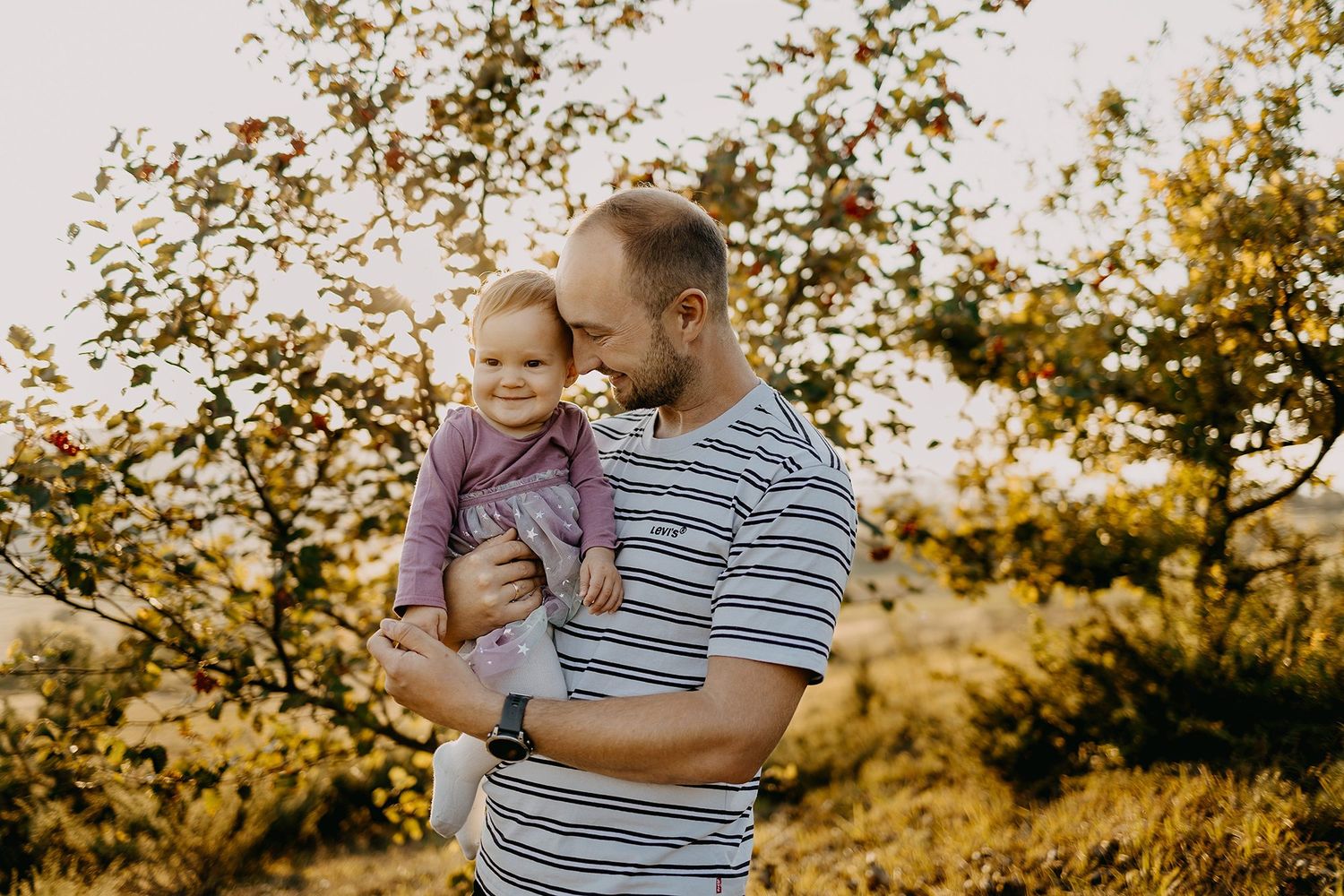 Father holding baby daughter in golden sunlight among autumn trees.