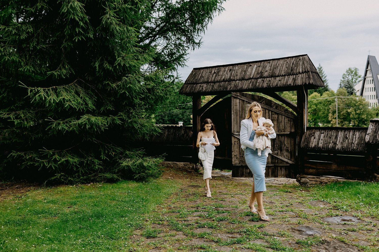 Two women walk through a garden gate, one carrying a baby after a christening ceremony.