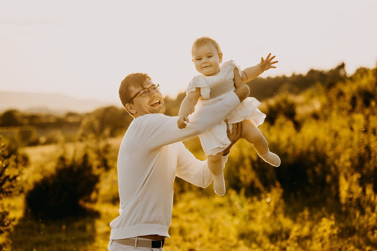 Father lifts laughing baby girl high in the air during golden hour outdoors.
