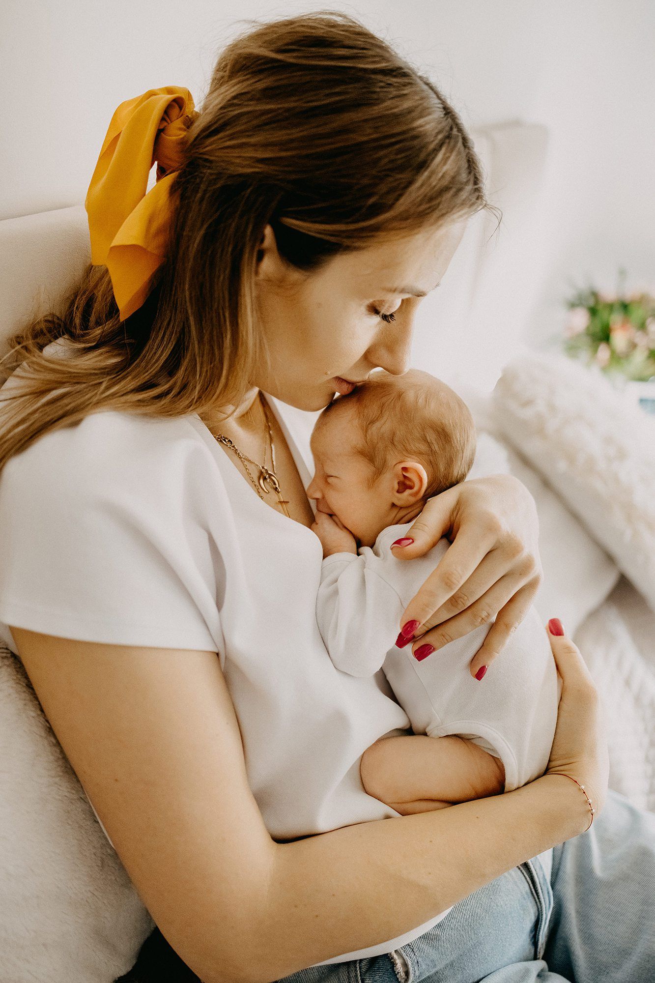 A mother with a yellow hair bow gently kisses her sleeping newborn baby indoors.