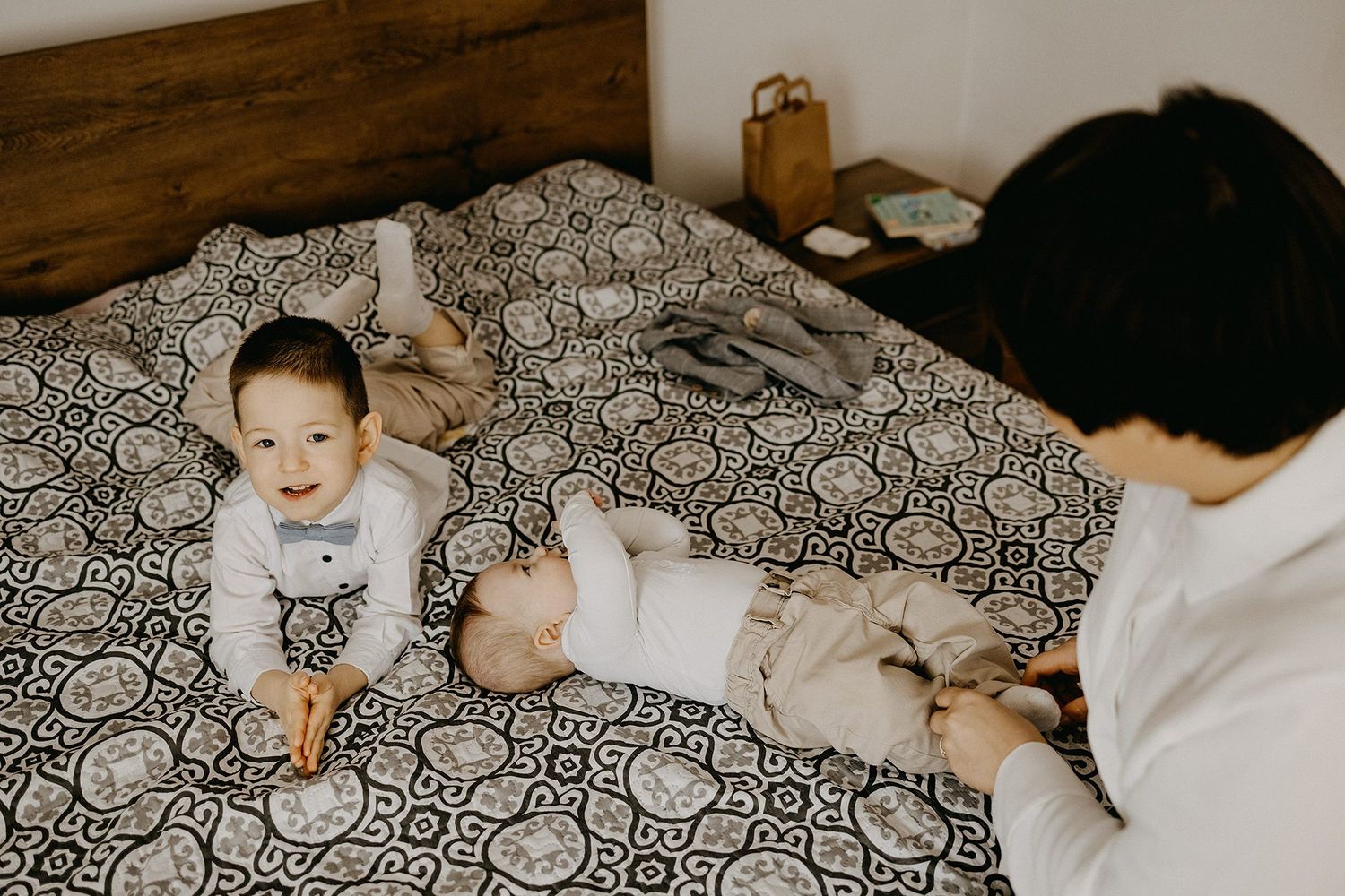 Two babies in formal attire lay on a patterned bed while a woman watches over them lovingly.