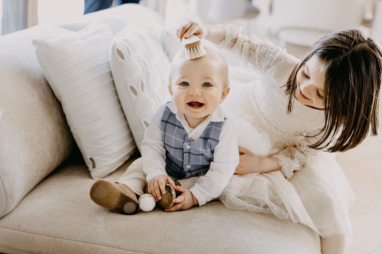 A mother playfully styles her laughing baby's hair on a cream-colored sofa.