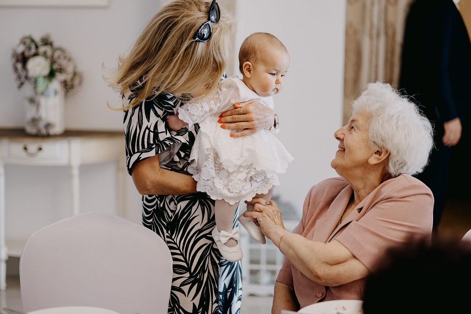 Elderly woman admiring a baby in a white christening dress held by a younger woman.