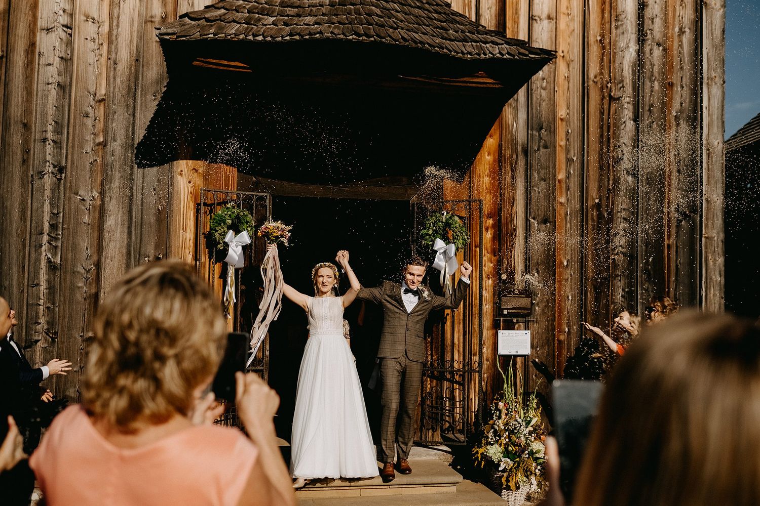 Joyful newlyweds raising arms in celebration as they exit a rustic wooden church.