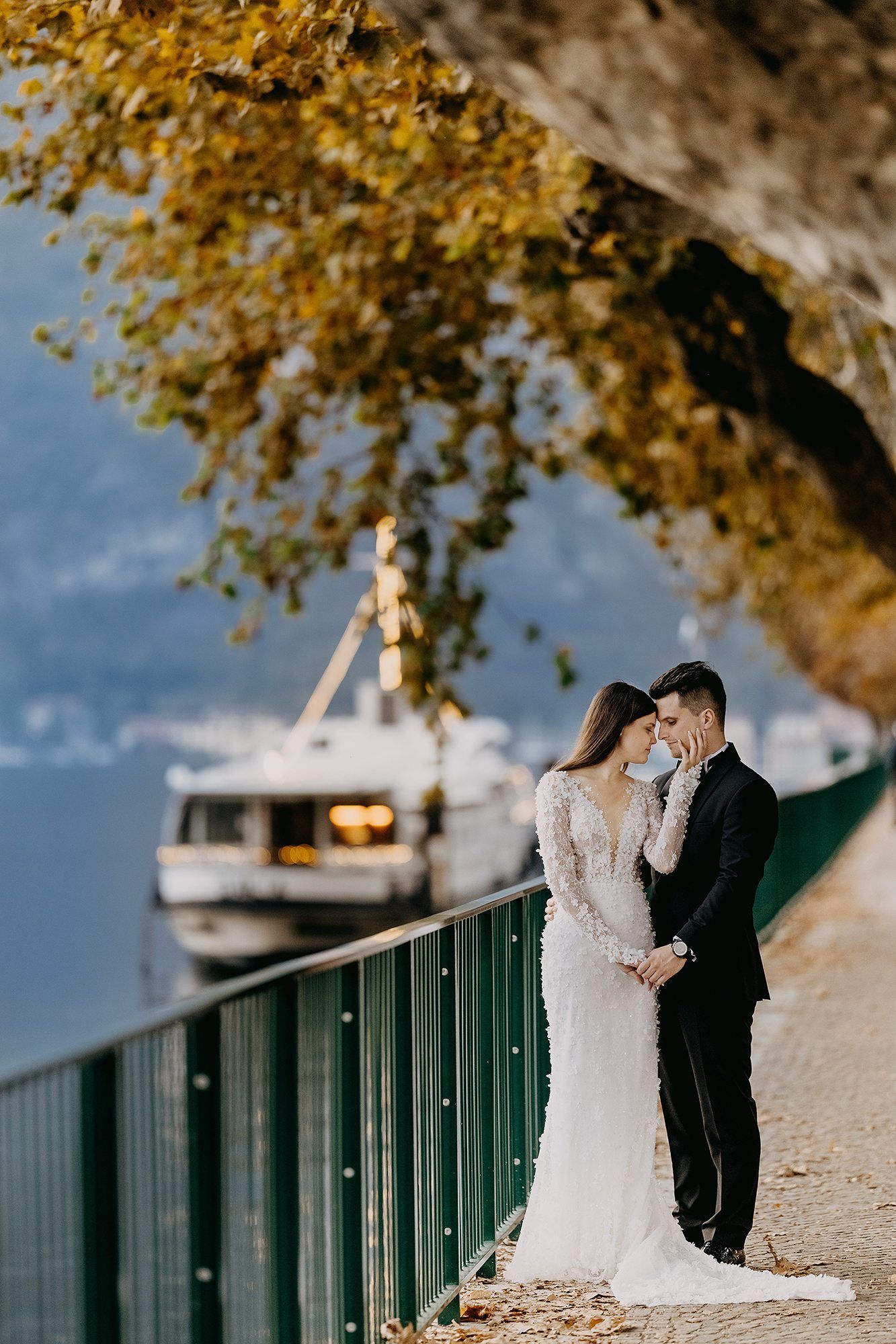 Bride and groom lean close under golden autumn foliage along a lakeside promenade with a yacht.
