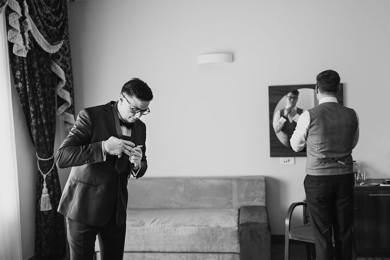 Groom adjusting cufflinks in black and white photo while assistant helps in mirrored reflection.
