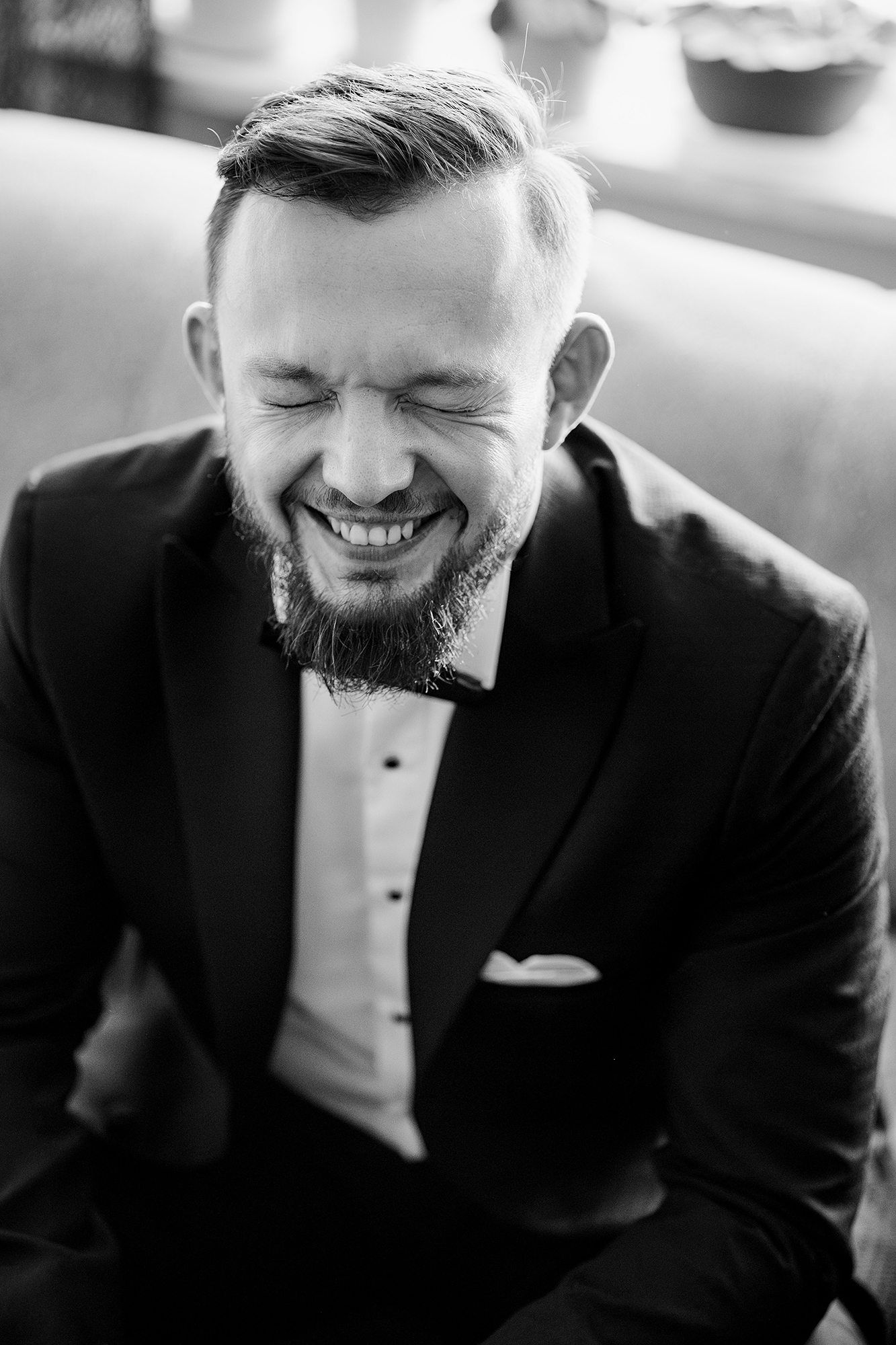 Joyful groom in black tuxedo and bow tie laughing in black and white portrait.