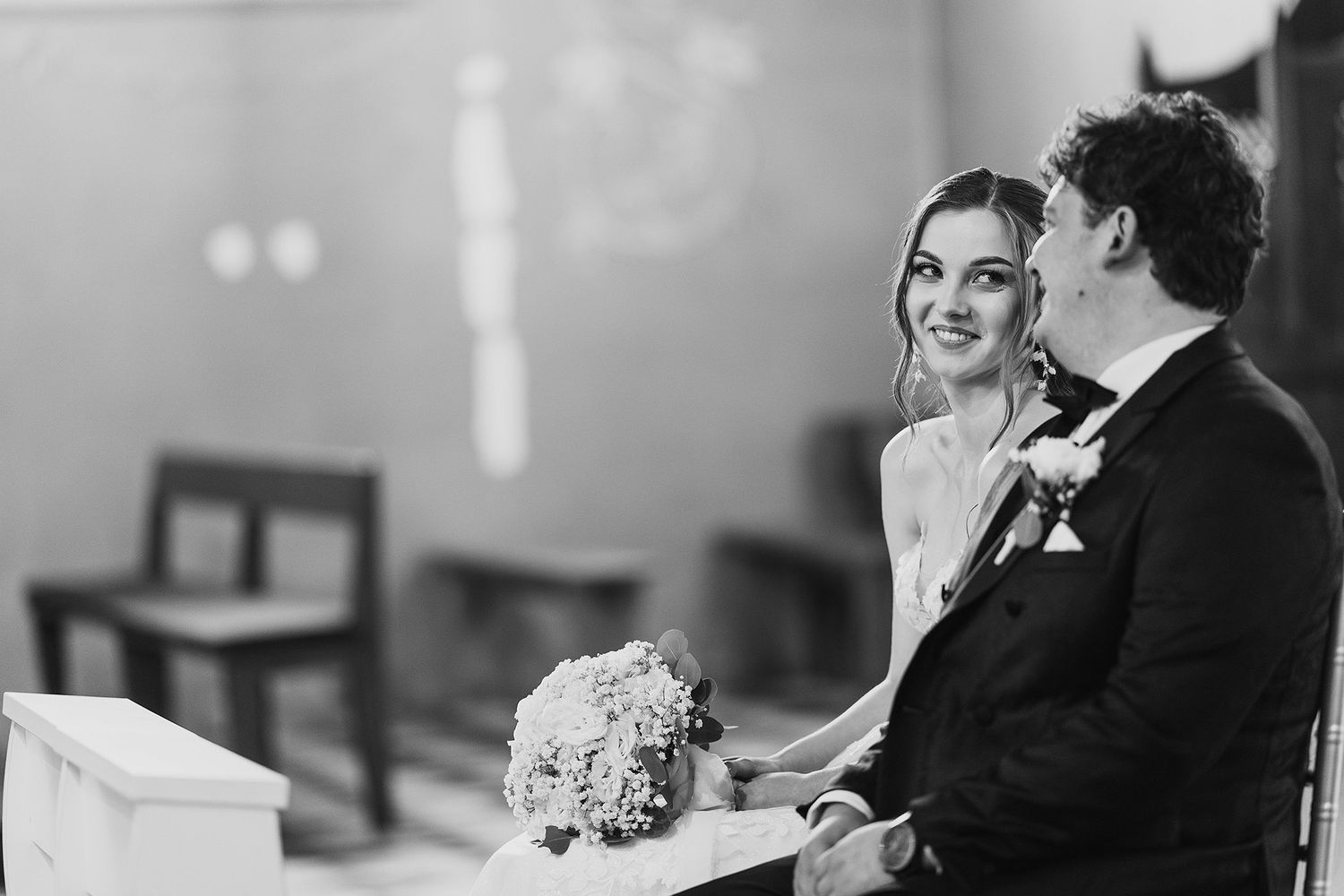 Black and white photo of smiling bride holding bouquet seated beside groom during church ceremony.