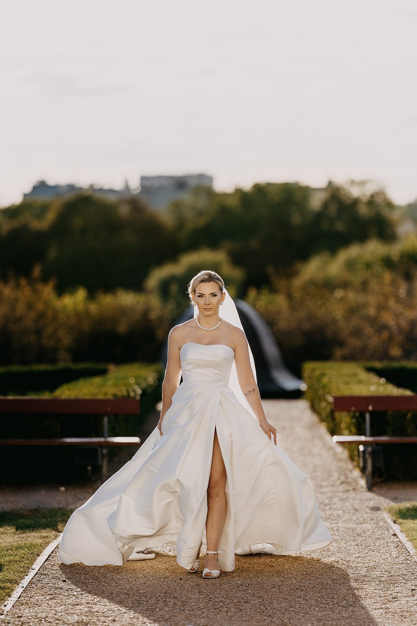 Confident bride in a strapless white gown with a slit posing in a formal garden path.