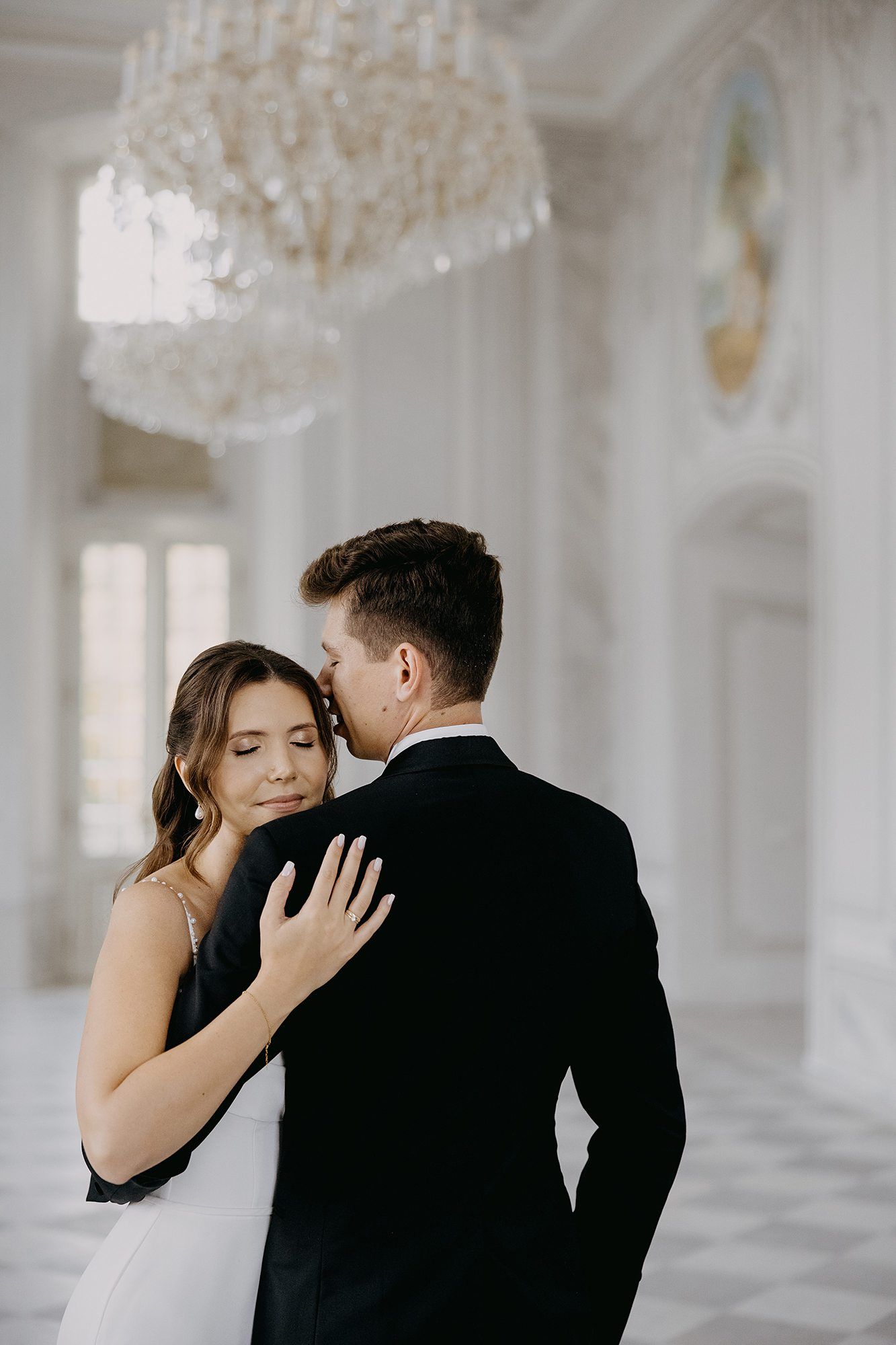 Groom tenderly kissing bride under a crystal chandelier in a grand white hall.