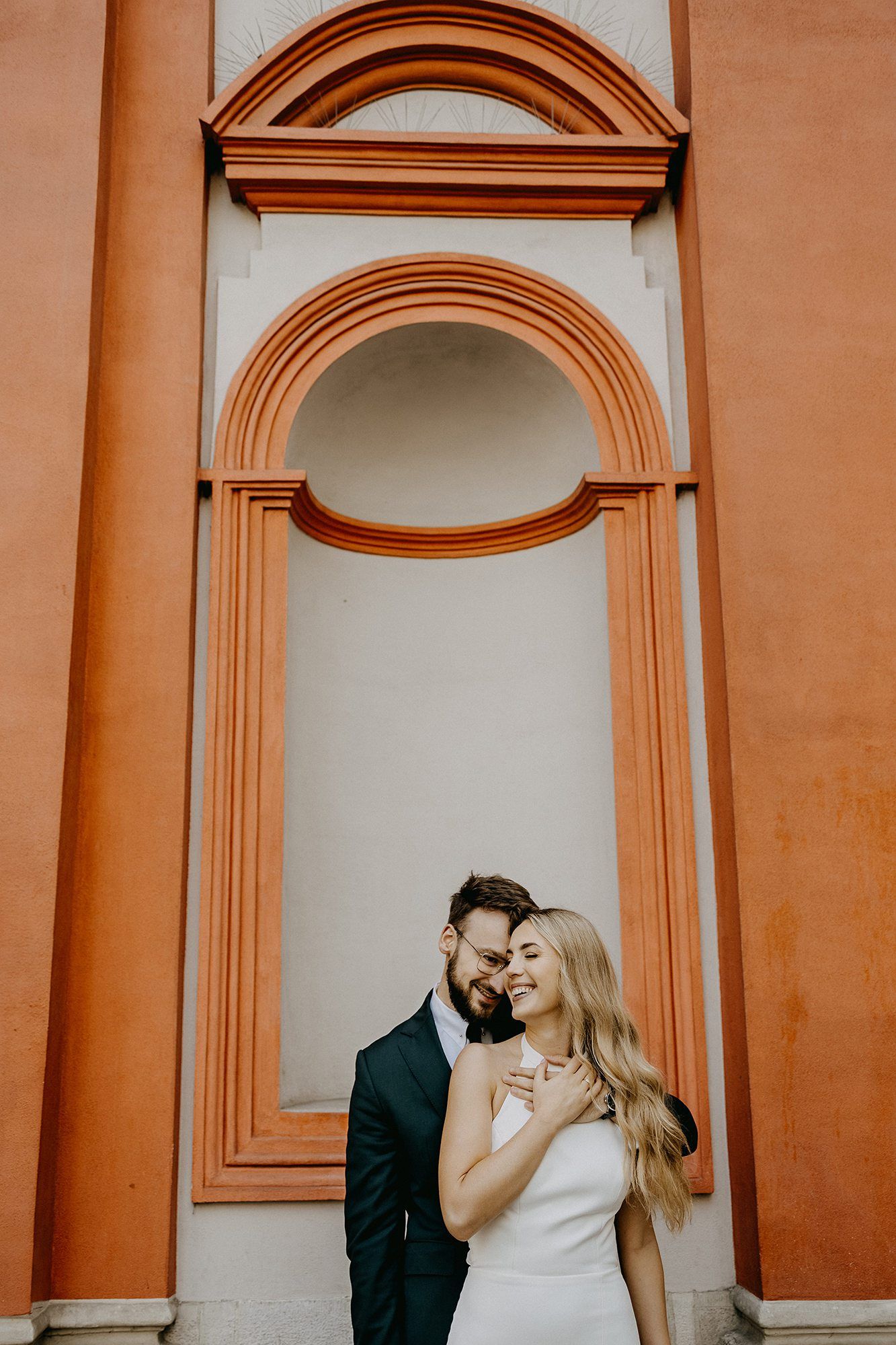 Bride and groom embracing before ornate terracotta arch on historic building.