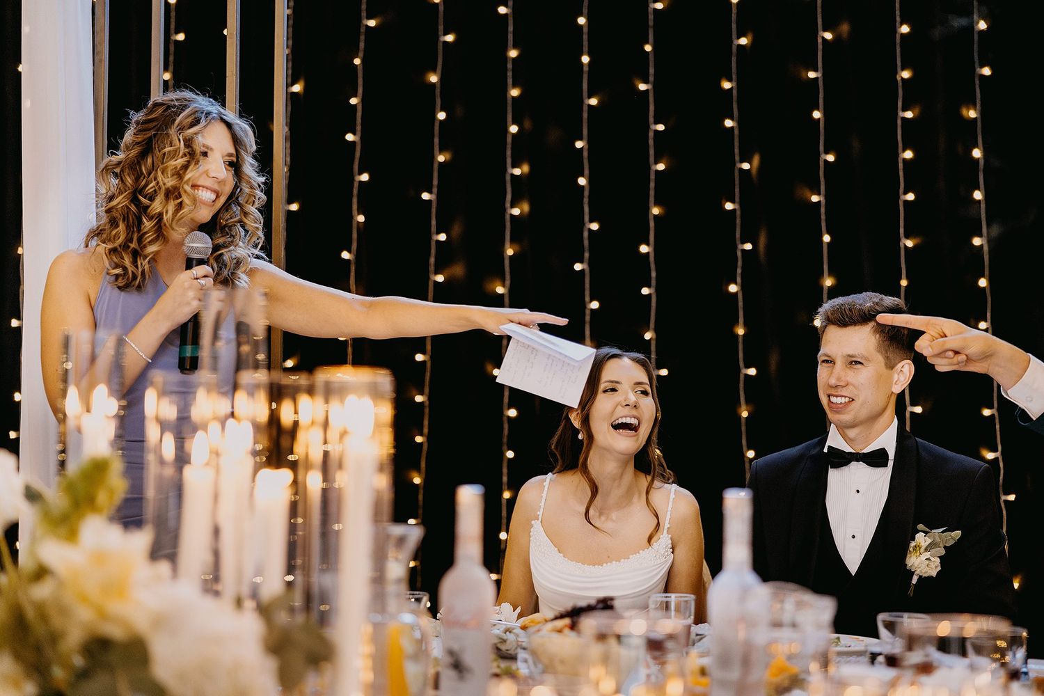 Wedding guest giving a toast while bride laughs joyfully at a candlelit reception table.