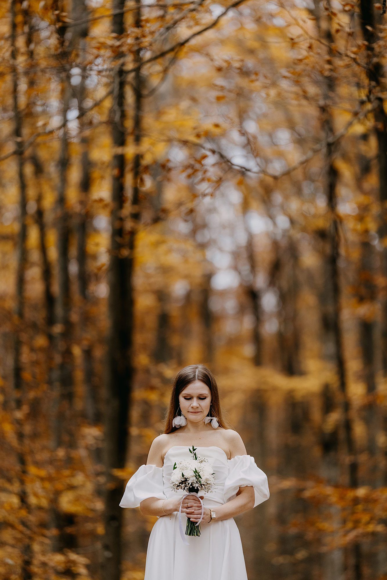 Elegant bride in off-shoulder white gown holds bouquet in golden autumn forest setting.