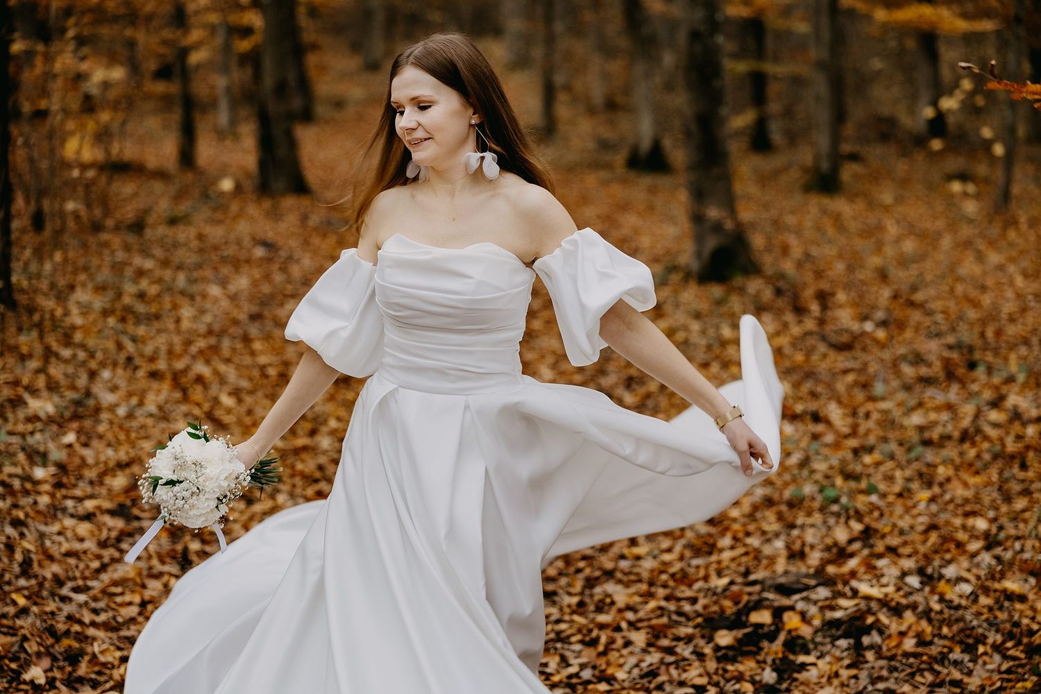 Bride twirls in white off-shoulder gown holding bouquet among golden autumn forest leaves.