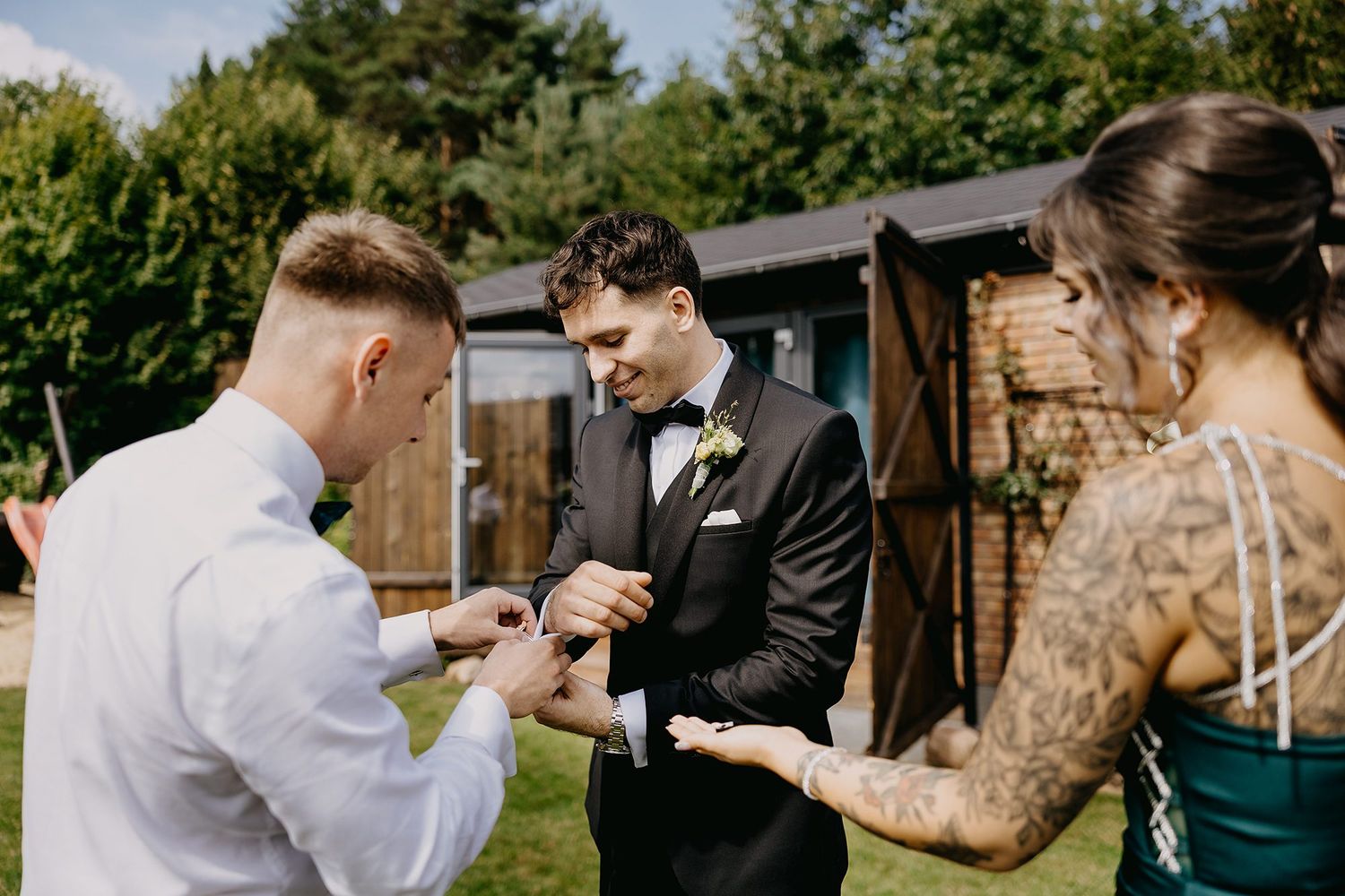 Groom being helped with his boutonniere by guests outdoors on a sunny wedding day.