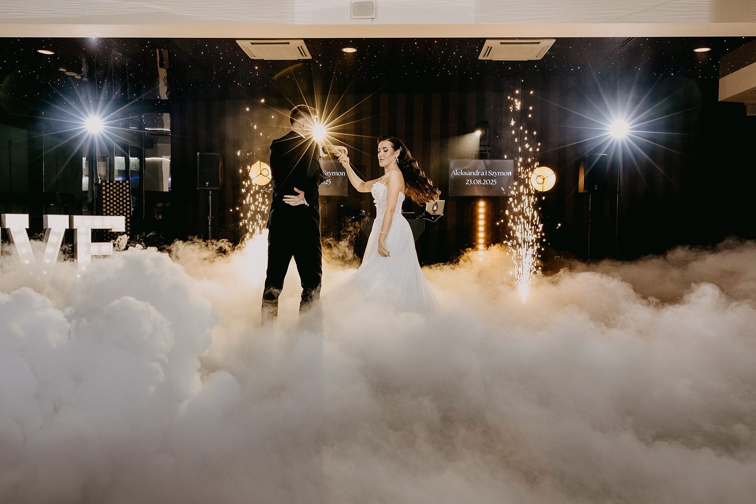 Bride and groom sharing first dance surrounded by dramatic dry ice fog and sparklers with a LOVE letter backdrop.
