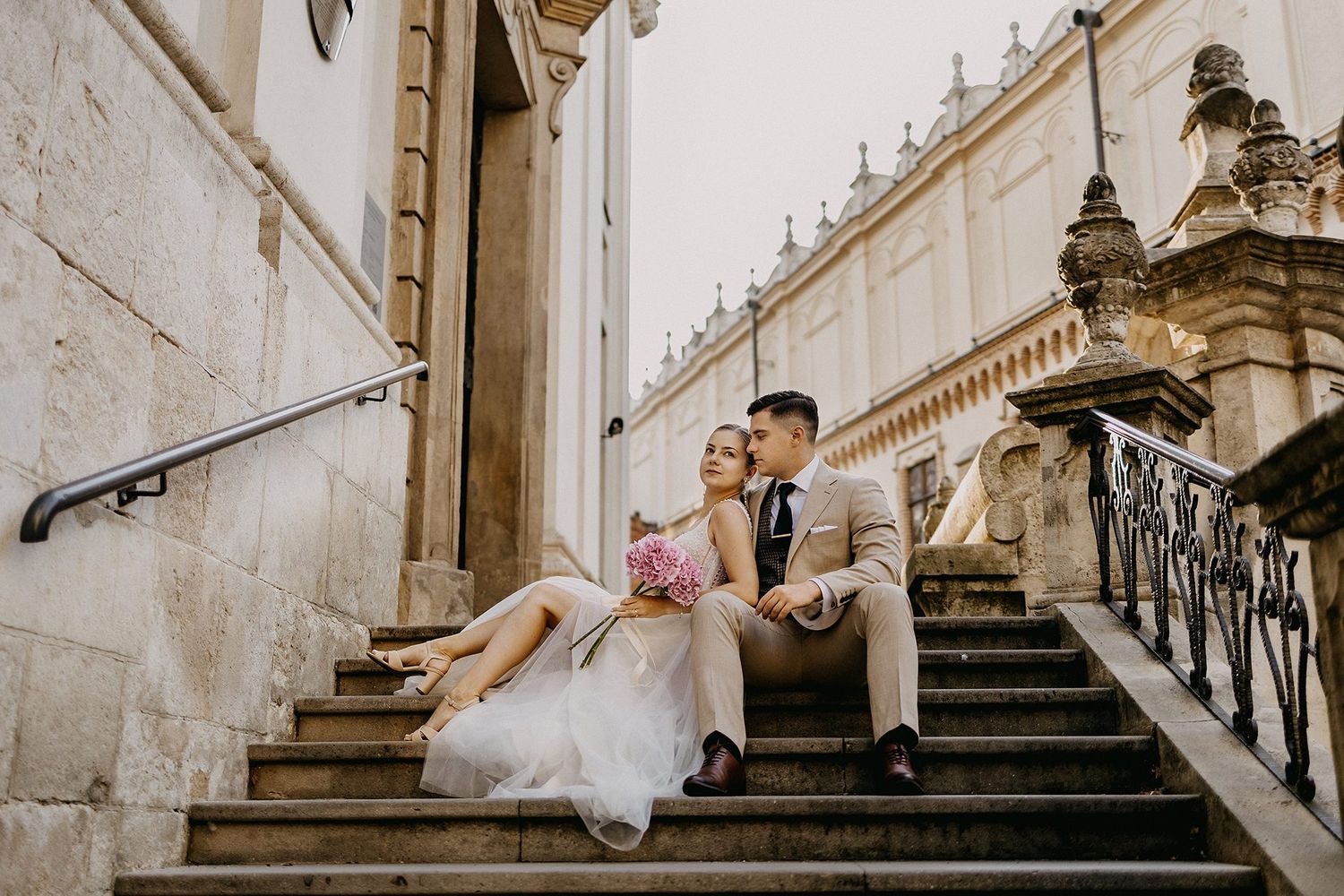 Wedding couple sits romantically on ornate stone steps of a historic baroque building.