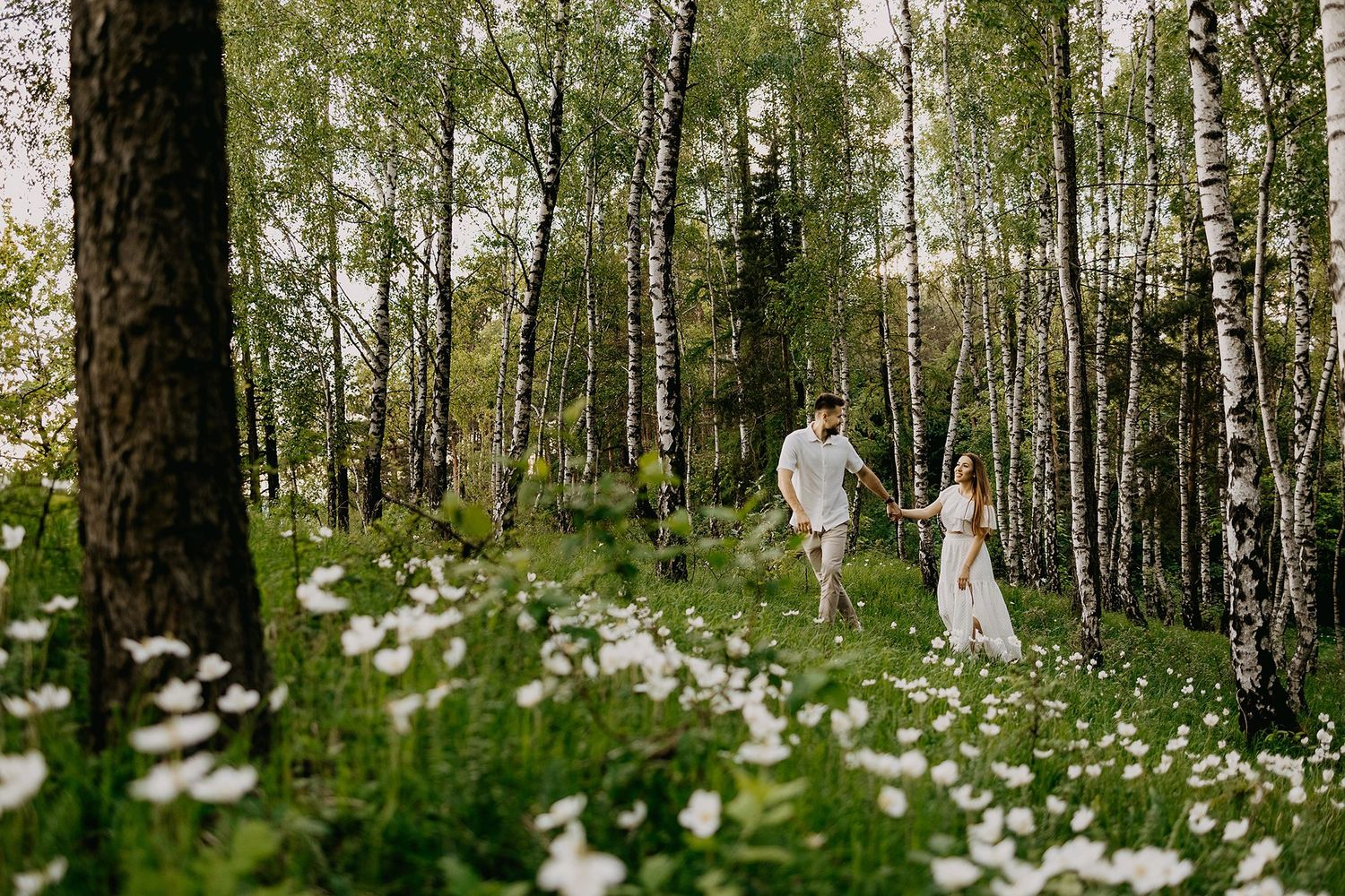 Couple holding hands walking through a birch forest with white wildflowers blooming on a sunny day.